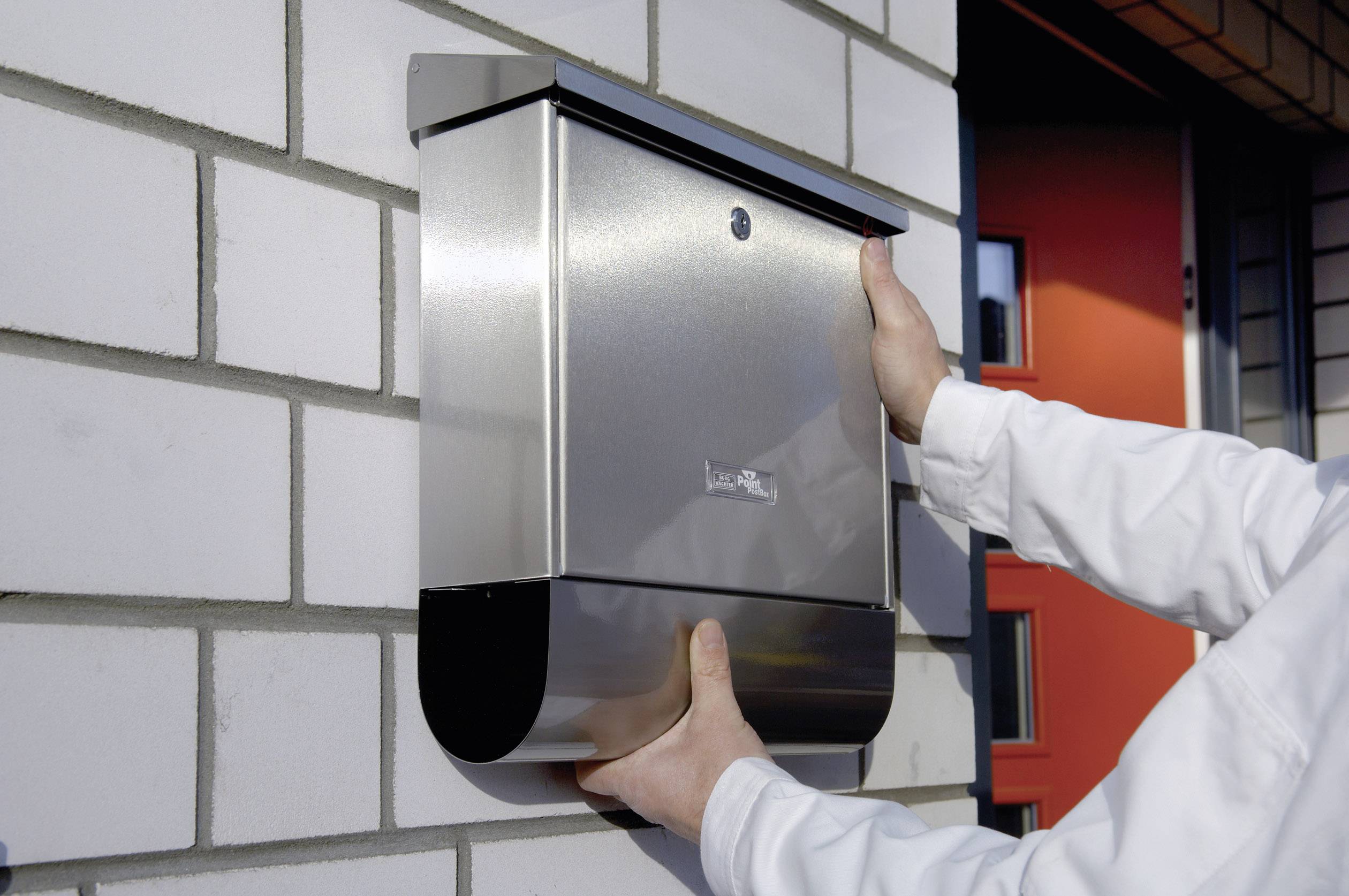 Egy személy egy ezüstszínű fémpostaládát szerel fel egy fehér téglafalra. A háttérben egy piros ajtó látható.