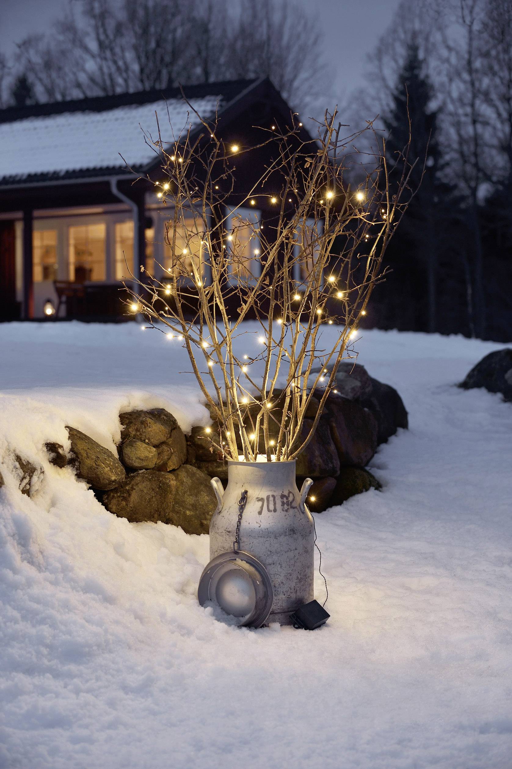 Egy díszített ág lámpácskákkal egy tejeskancsóban, havas kertben, kivilágított ház a háttérben.