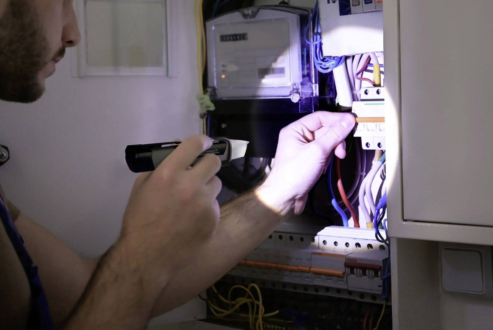 Egy személy egy zseblámpával egy elektromos elosztószekrényt vizsgál, amely tele van kábelcsatlakozásokkal.