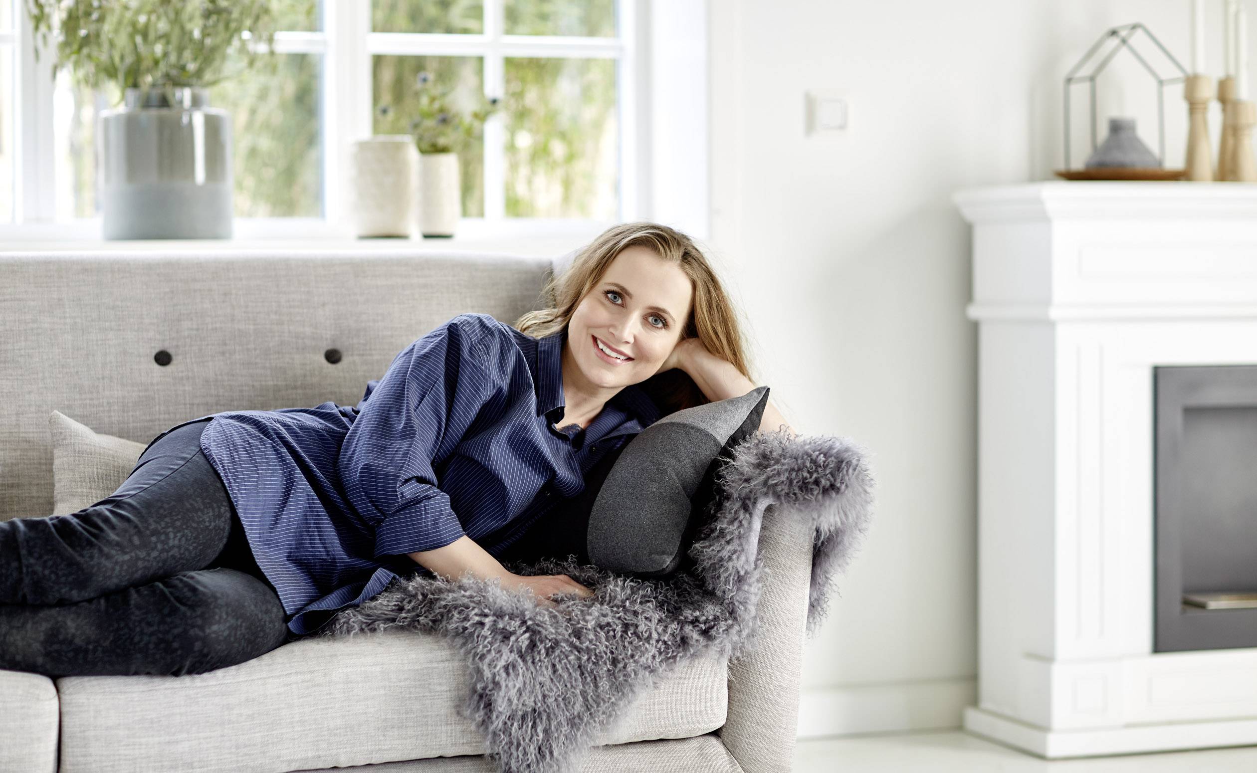 Egy személy lazán hever egy kanapén egy világos nappaliban. A háttérben ablakok és egy kandalló látható.