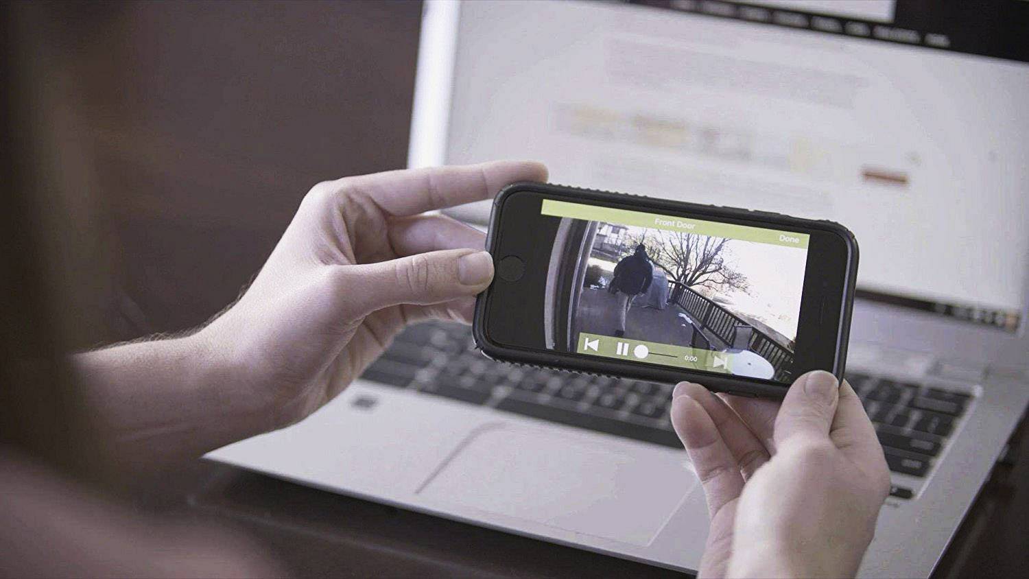 Egy személy egy laptoppal előtt tart egy okostelefont. A telefon képernyőjén egy erkély biztonsági kamerájának képe látható.