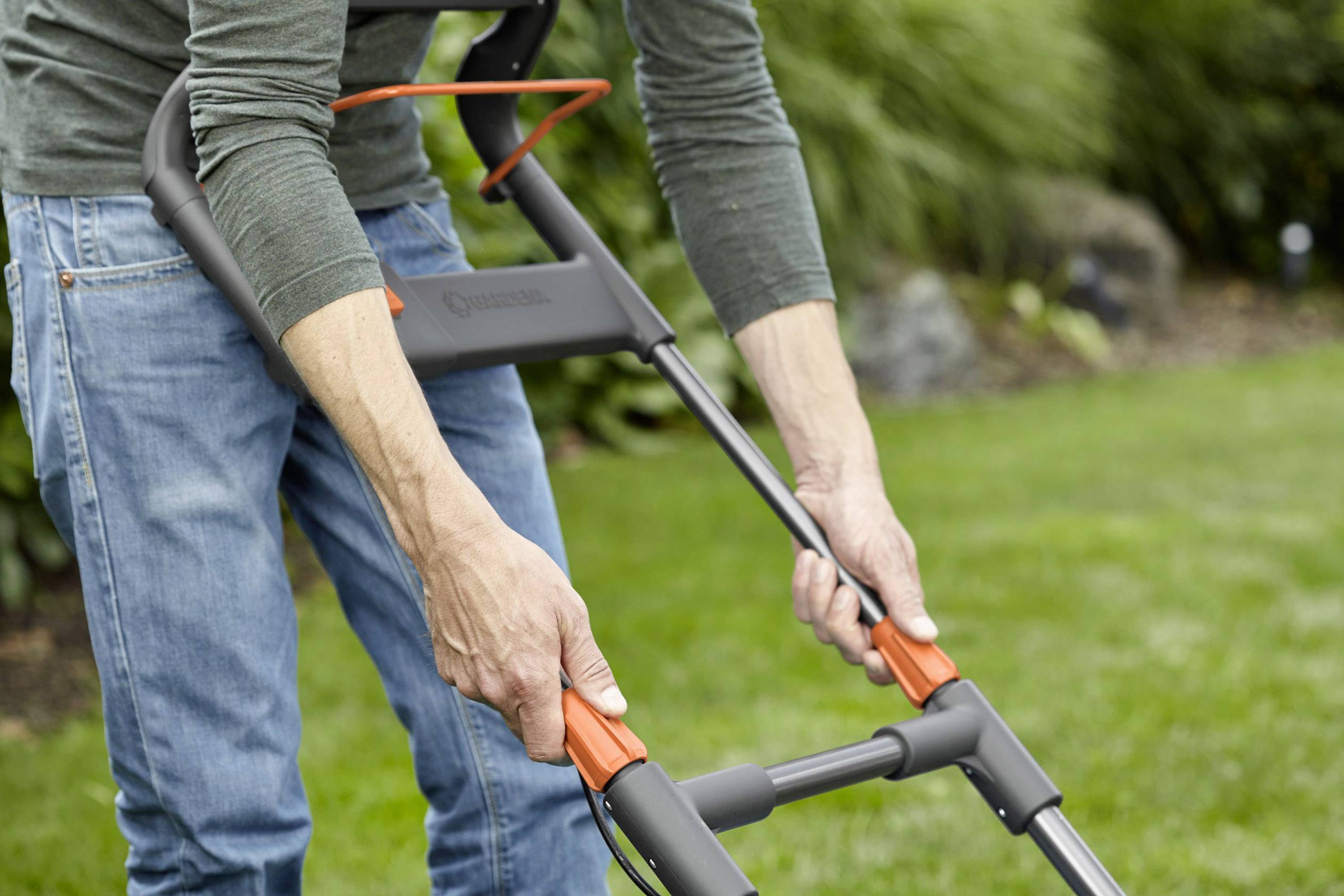 Egy farmer nadrágot viselő személy egy fűnyírógépet kezel egy kertben. Szorosan fogja a fogantyút, és a gyep rendezett.