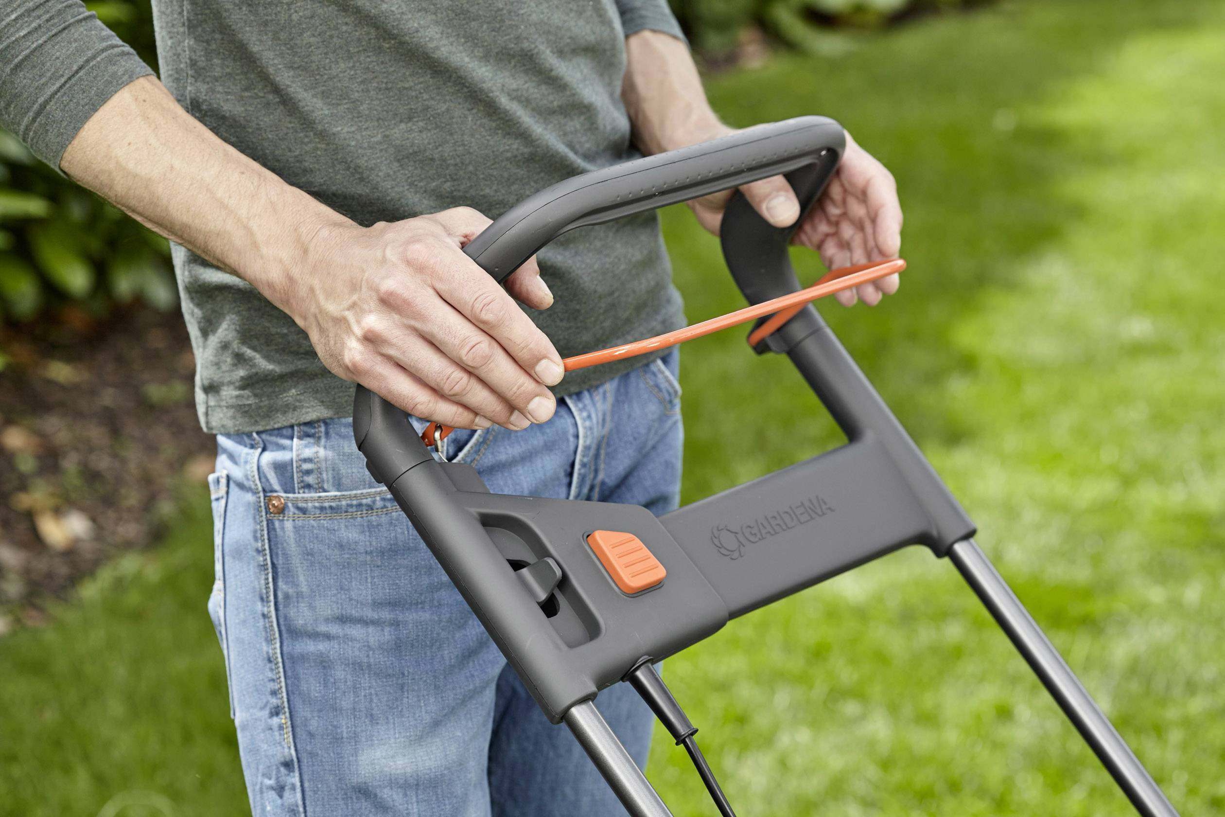 Egy ember fűnyírót tol egy zöld gyepen. Kezei a fogantyút fogják. A gyep gondozott, a háttérben bokrok láthatók.