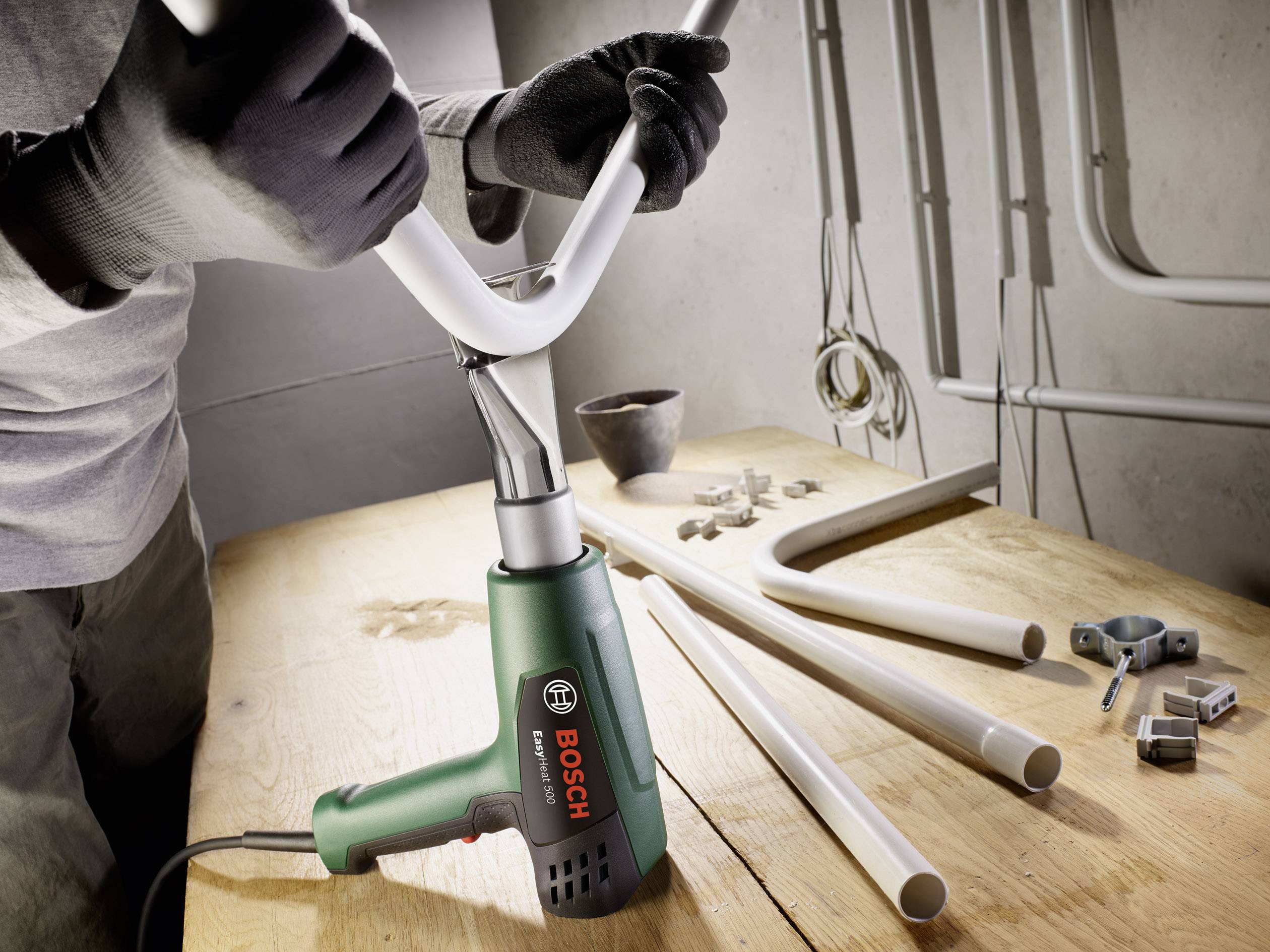 Egy személy egy Bosch hőlégfúvóval egy fehér csövet hajlít egy faasztalon, körülvéve további csövekkel és tartozékokkal.