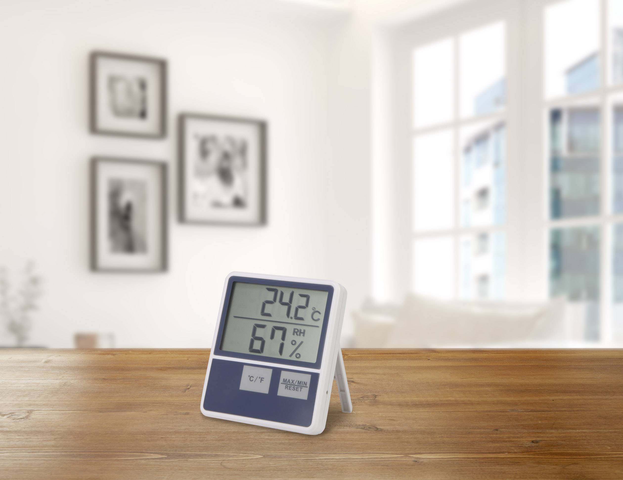 Egy digitális termo-higrométer egy faasztalon 24,2°C-ot és 67% páratartalmat mutat. A háttérben elmosódott ablakok és képek láthatók.