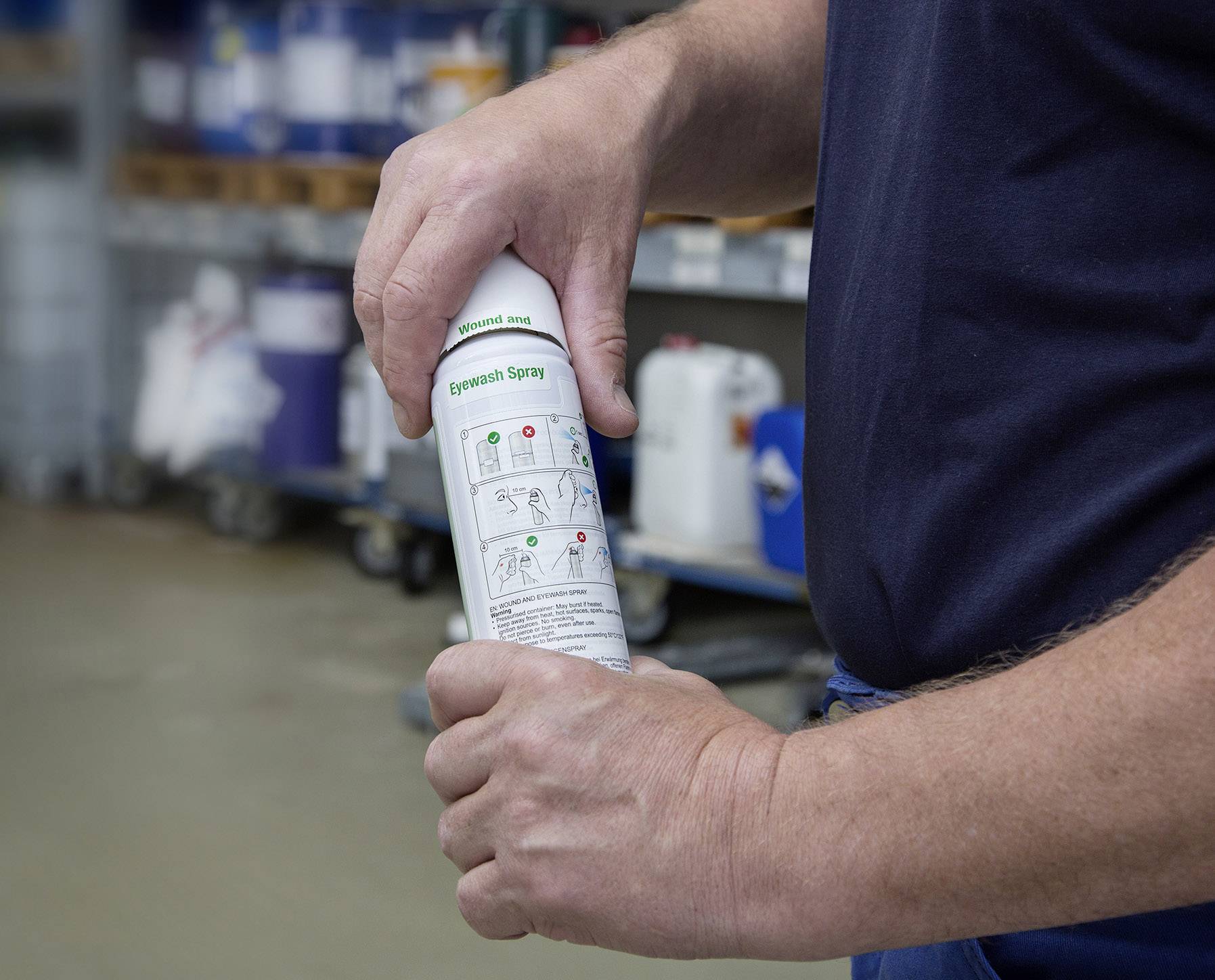 Egy szemöblítő spray-t tart egy személy, használatra készen. A háttérben polcok láthatók különböző tartályokkal.