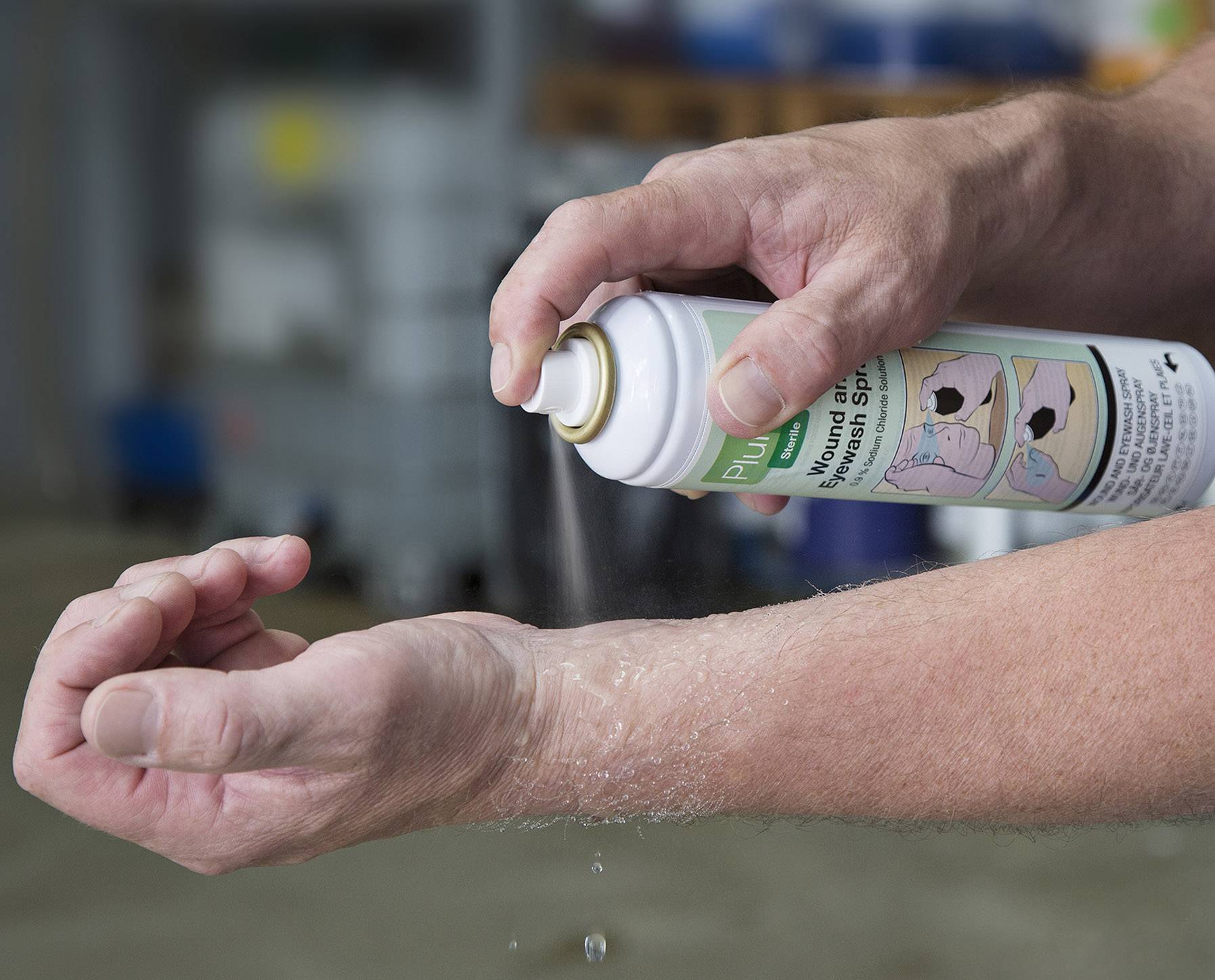 Egy személy elsősegély sprayt permetez a karjára, hogy kezeljen egy sérülést vagy irritációt. A háttérben egy elmosódott műhely látható.