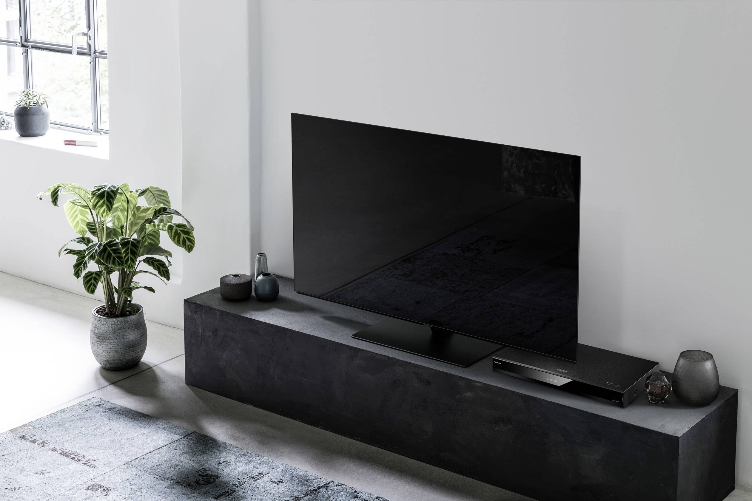 Egy modern televízió egy fekete TV-asztalon áll egy zöld szobanövény mellett, egy világos szobában, szőnyeggel.