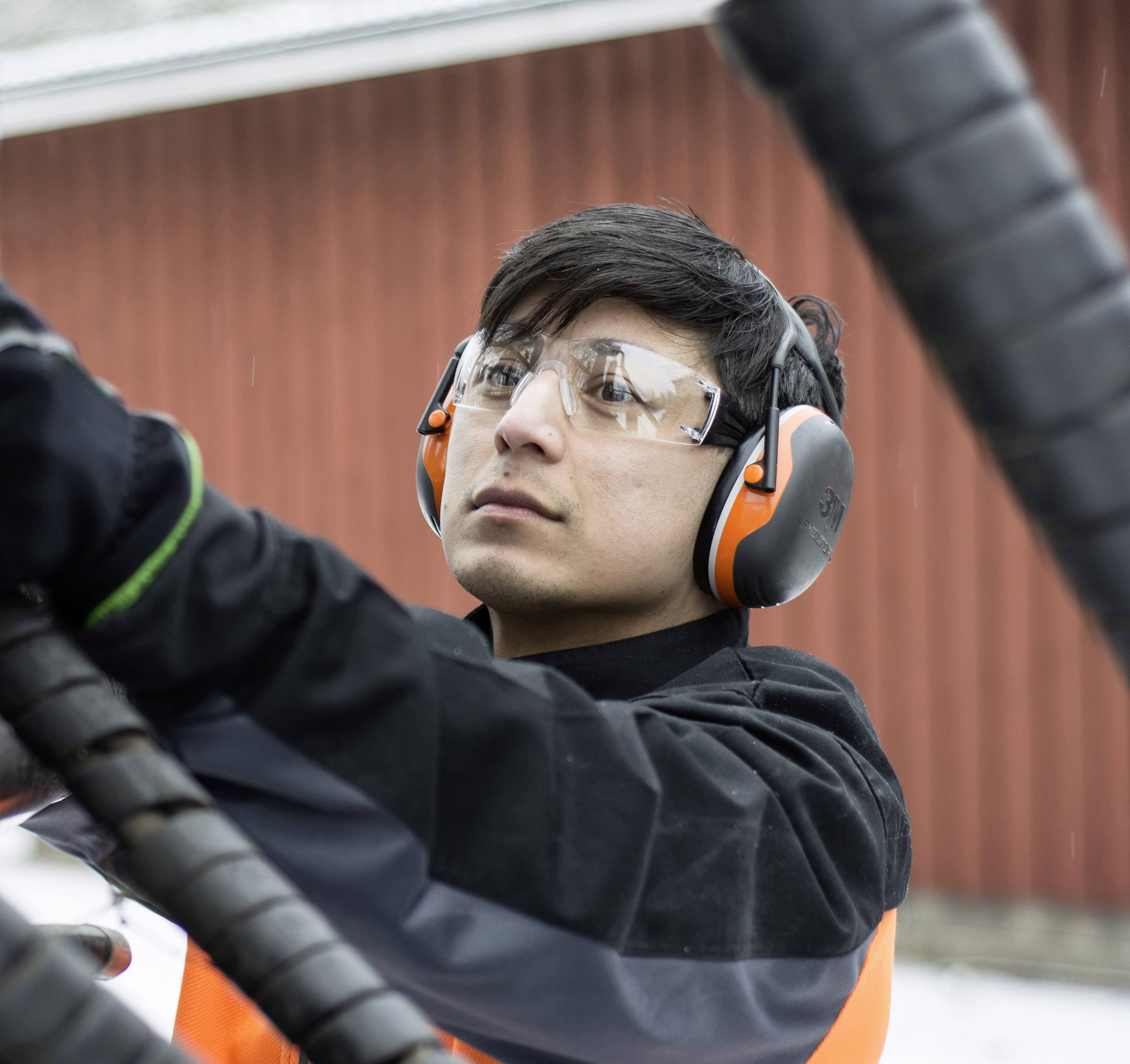 Egy személy védőszemüveget és fülvédőt visel, és egy gépen dolgozik a szabadban. A háttérben egy piros épület látható.