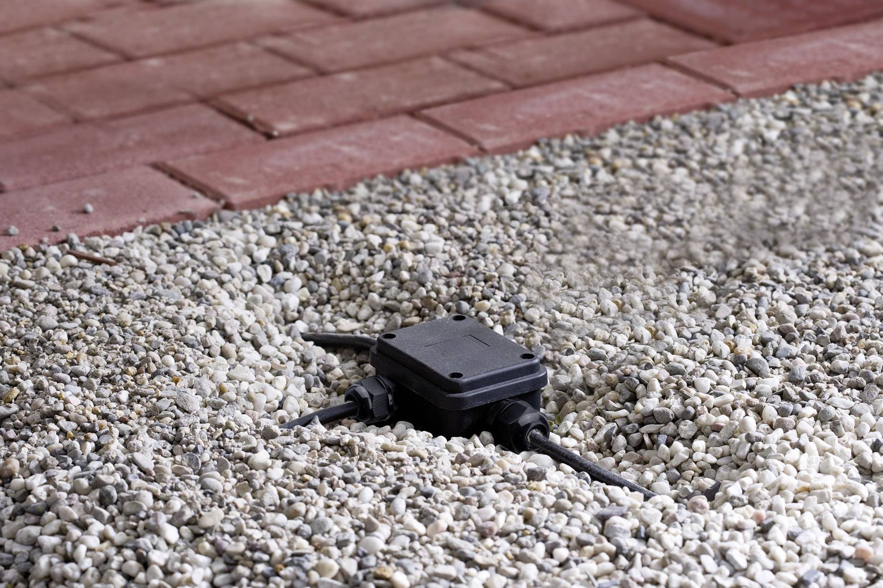 Egy fekete elektronikai doboz fekszik a kavicson egy térköves út mellett. Kábelek futnak a doboz mindkét oldalán.