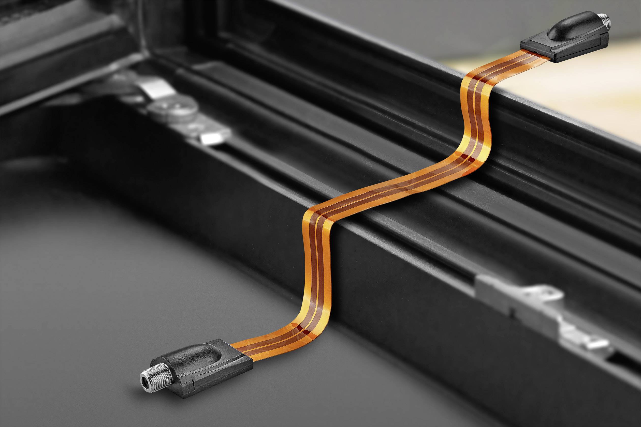 Egy lapos kábel húzódik egy ablak és kerete között, bemutatva a kábel rugalmasságát és alkalmazkodóképességét szűk terekben.