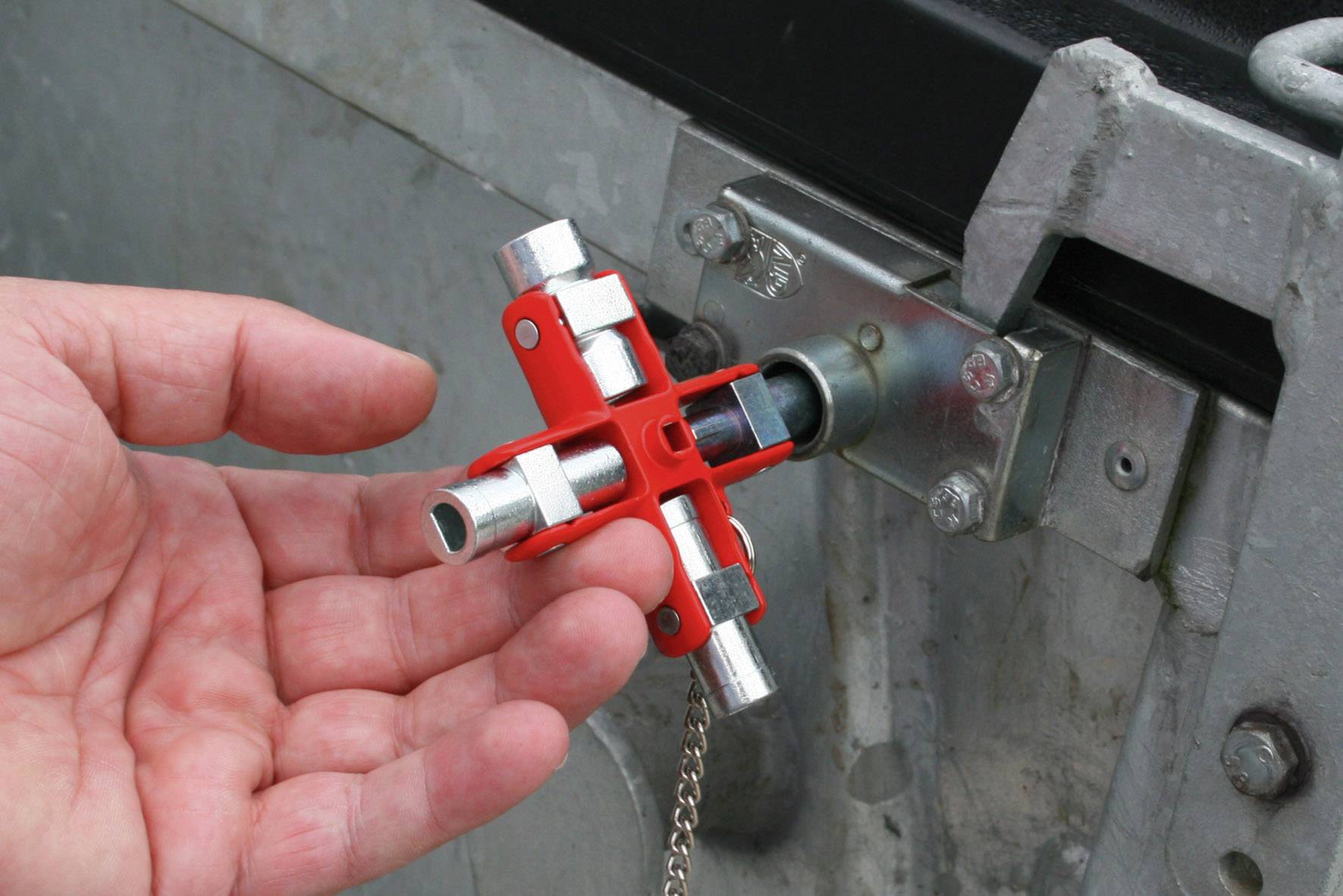 Egy kéz egy piros négyélű kulcsot tart egy fémkapu zárszerkezeténél. A kulcs különböző méretű fogakkal rendelkezik a zár kinyitásához.