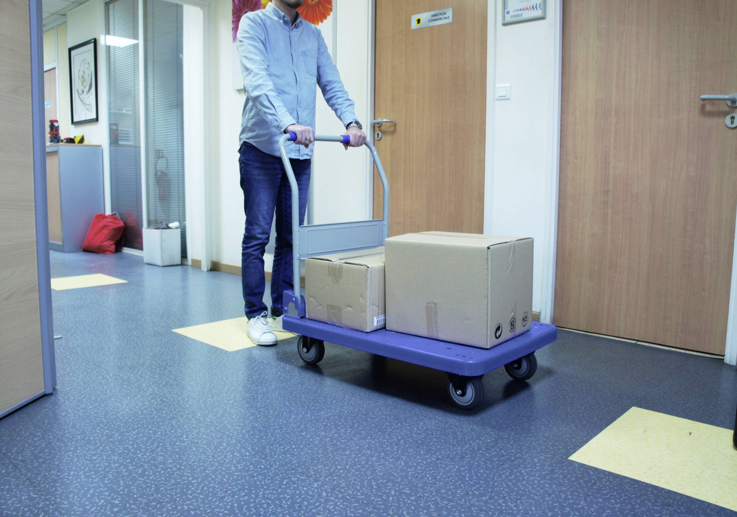 Egy személy két nagy dobozos targoncát tol egy irodafolyosón. A háttérben ajtók és irodai kellékek láthatók.