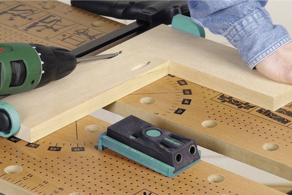 Kéz tart egy fadarabot a munkapadon; elektromos fúrógép készül lyukat fúrni. A munkapadon lévő jelölések segítenek a precíz munkavégzésben.