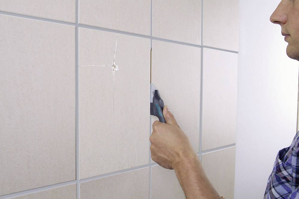 Egy személy egy csempézett falról egy törött csempét távolít el egy eszköz segítségével, valószínűleg azért, hogy megjavítsa vagy kicserélje.