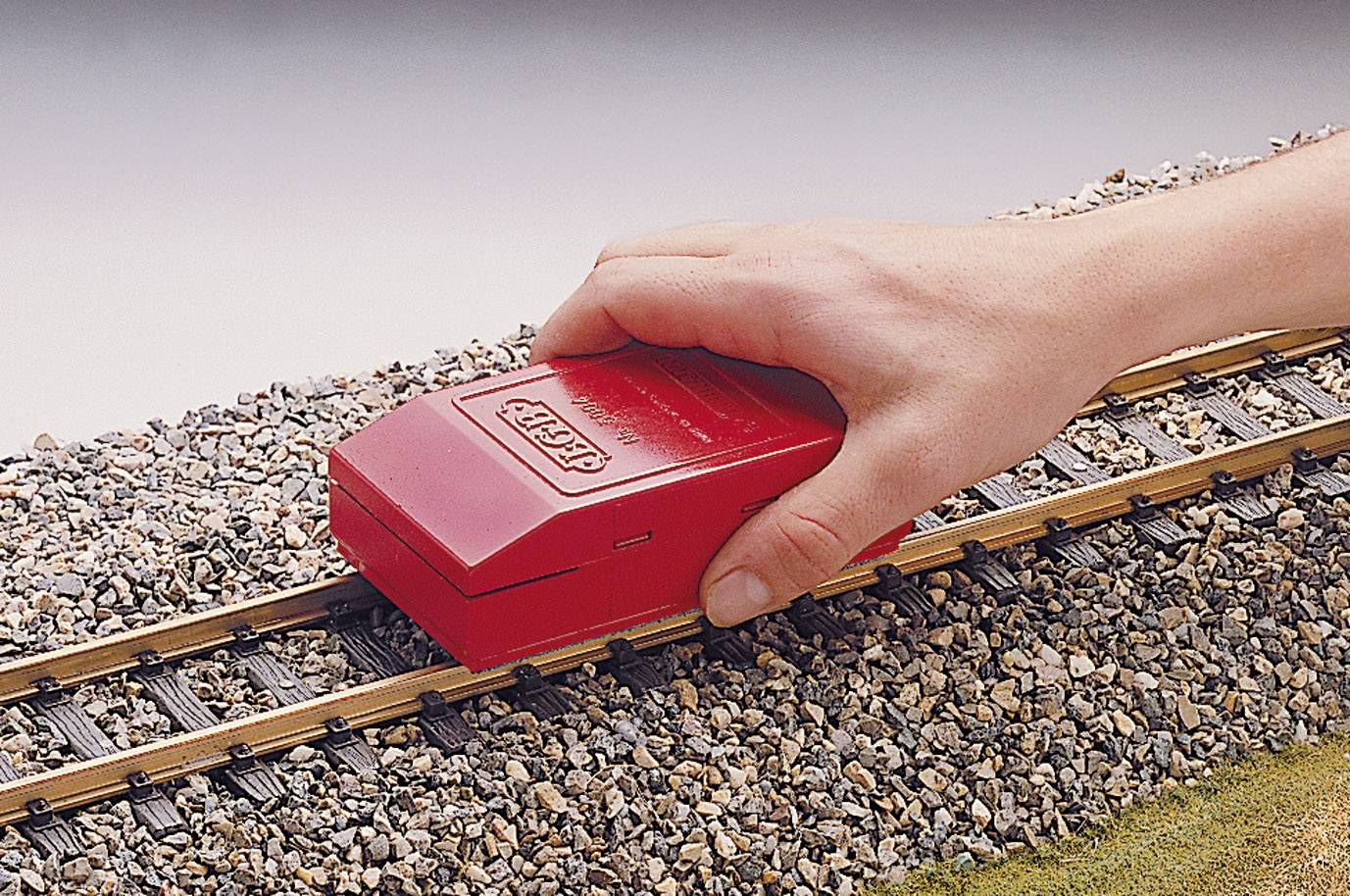 Egy kéz egy piros síntisztító készüléket tart egy modellvasúti sínpályán, kavicságyon, amelyet a sínek tisztítására használnak.