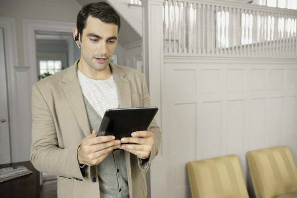 Egy férfi bézs zakóban egy tabletet használ egy modern irodában, bluetooth-os headset van a fülén. A háttérben világos falak és székek láthatók.