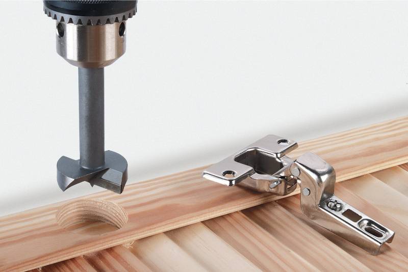 Egy Forstner-fúró lyukat fúr egy falemezbe, mellette egy zsanér fekszik. Téma: Bútorszerelés.