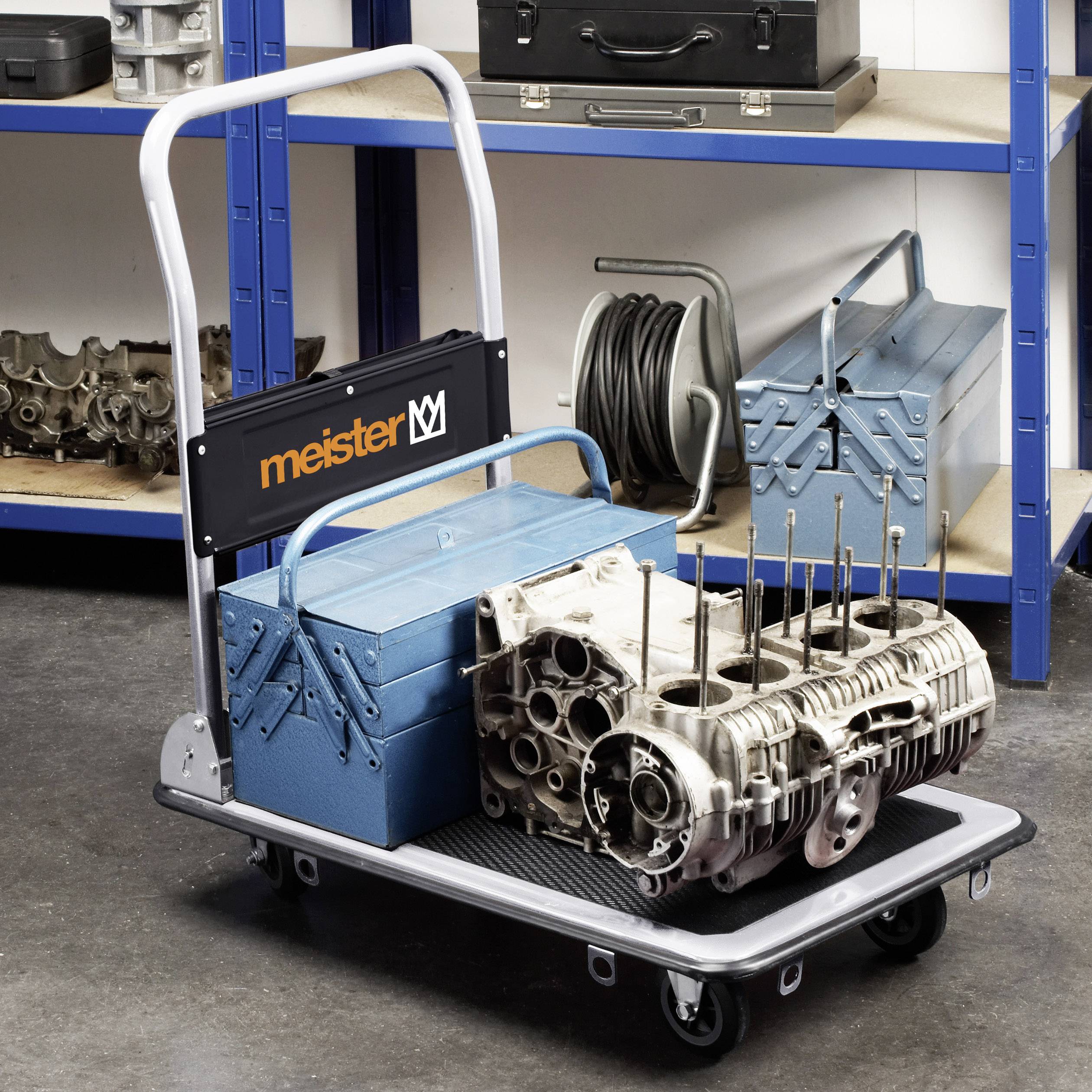 Műhelykocsi 'mester'-logóval, rajta kék fémláda és motorrész hengerrel, a háttérben szerszámospolicok és kábelek.