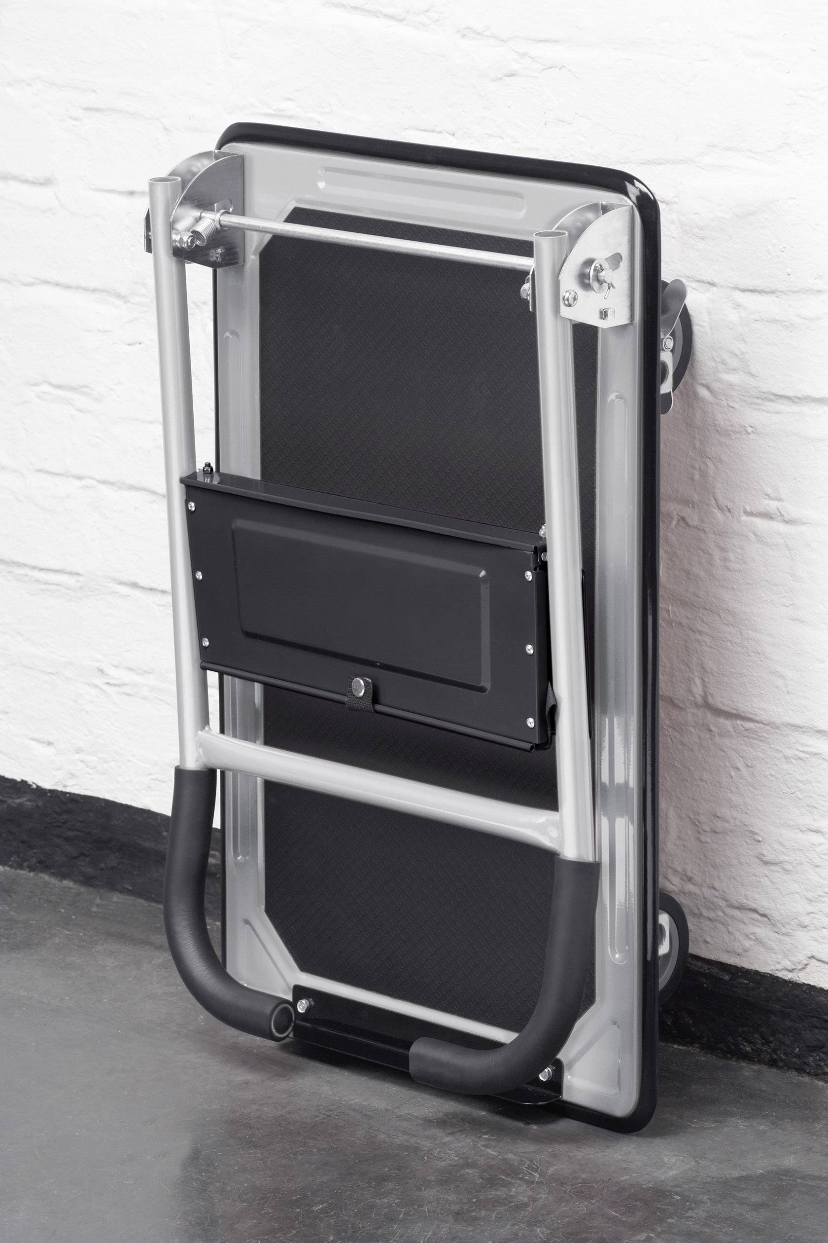 Egy felhajtott kézikocsi egy fehér téglafal mellett áll. A fekete és ezüst kocsi össze van csukva és helytakarékosan elhelyezve.