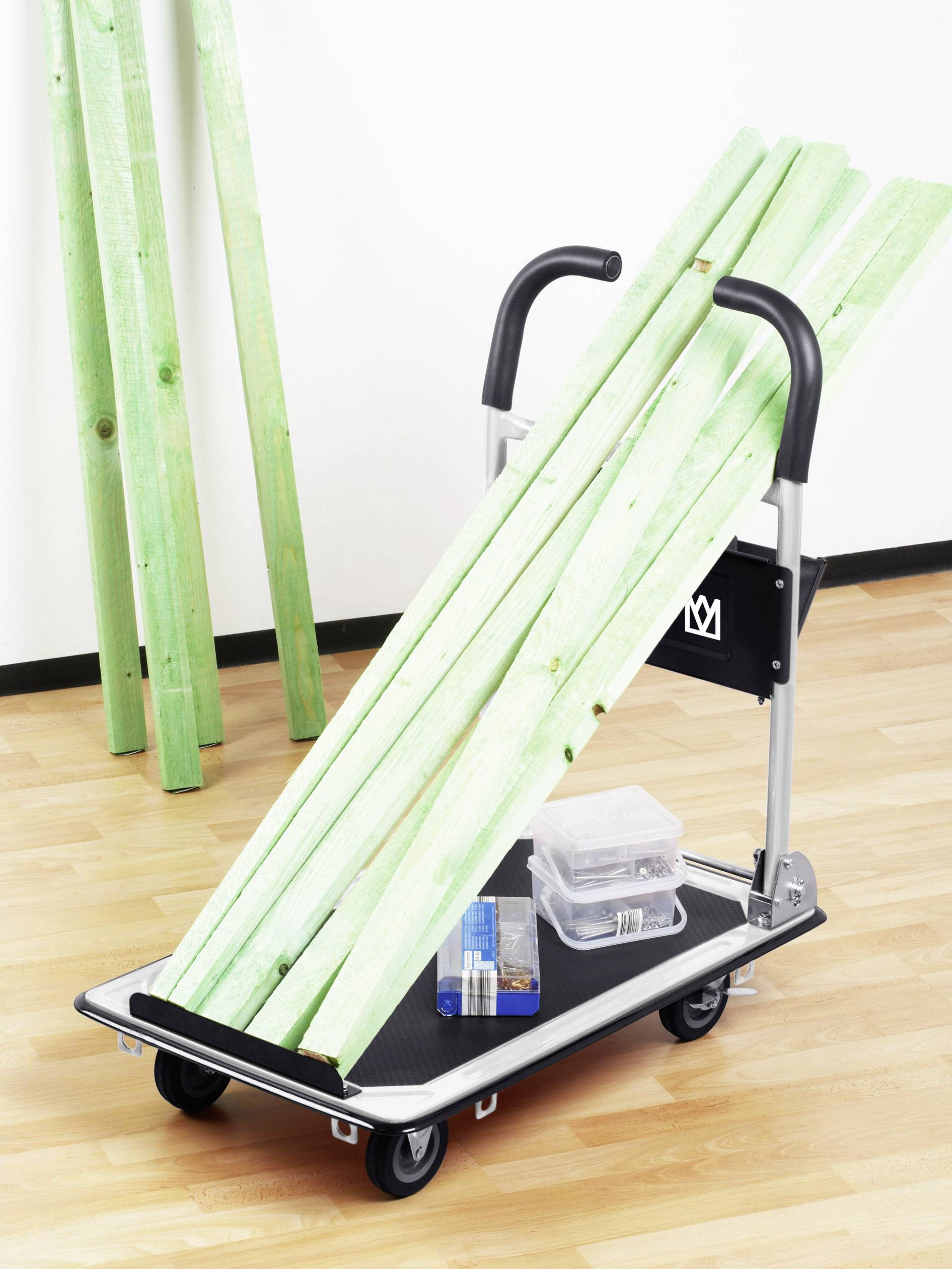 Egy fémből készült talicska fa padlón zöld faléceleket és egy műanyag dobozt szállít szerszámokkal. A háttérben lécelő rakatok vannak a fal mellett.