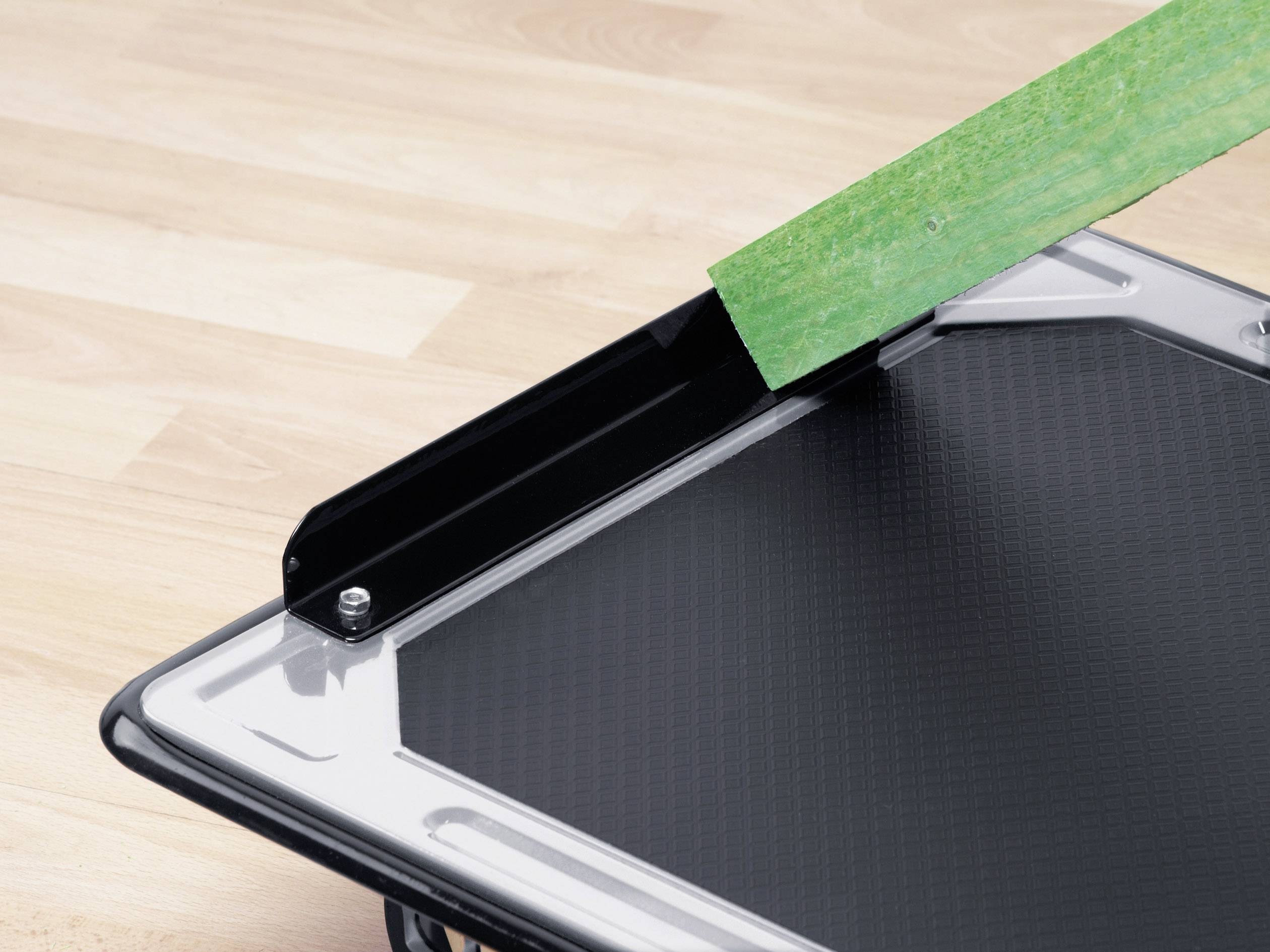 Egy fekete vágódeszka egy pengével, amely egy zöld anyagot vág egy faasztalon.