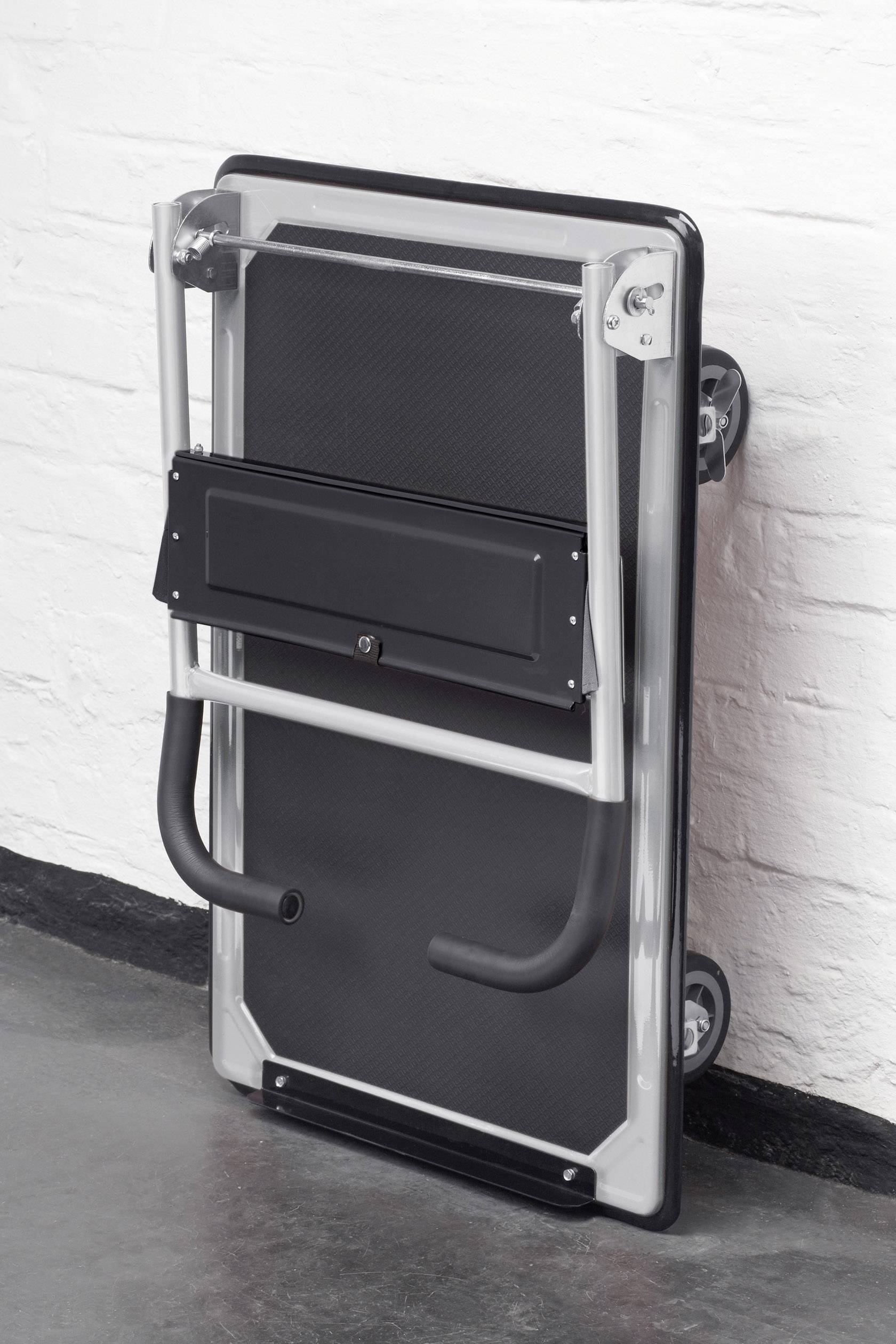 Összecsukott asztal görgőkkel egy fehér téglafalnak támasztva, tárolásra vagy szállításra kész állapotban.