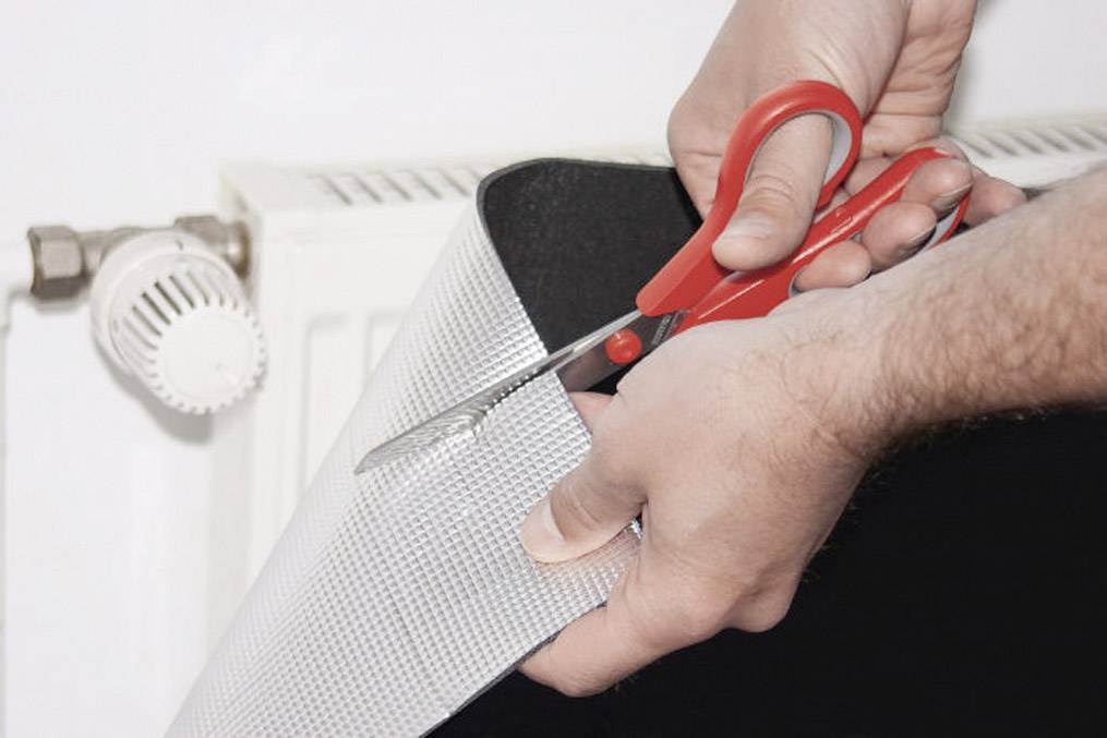 Egy személy egy piros ollóval vágja a fényvisszaverő fóliát egy fehér radiátor közelében, hogy energiát takarítson meg.