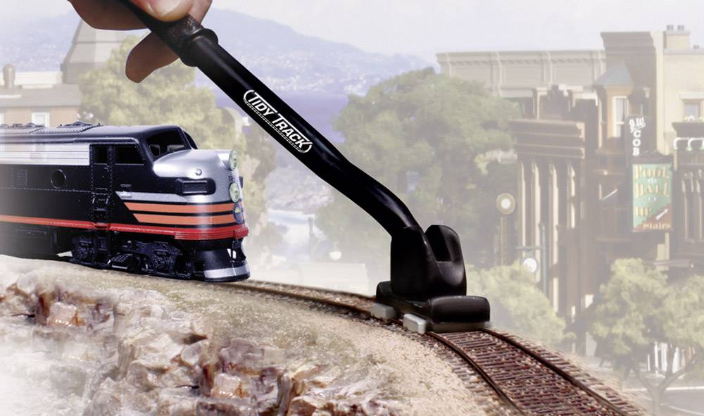 Egy kéz egy eszközzel tisztítja egy modellvasút sínét egy tájképben, ahol hegyek és épületek láthatók a háttérben.