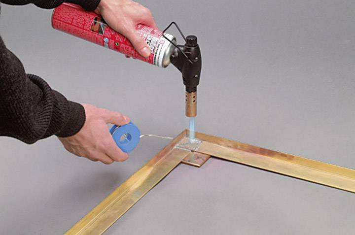 Két kéz, amint egy forrasztólámpát és forrasztóónt használ két fémdarab sarkának összekötéséhez.