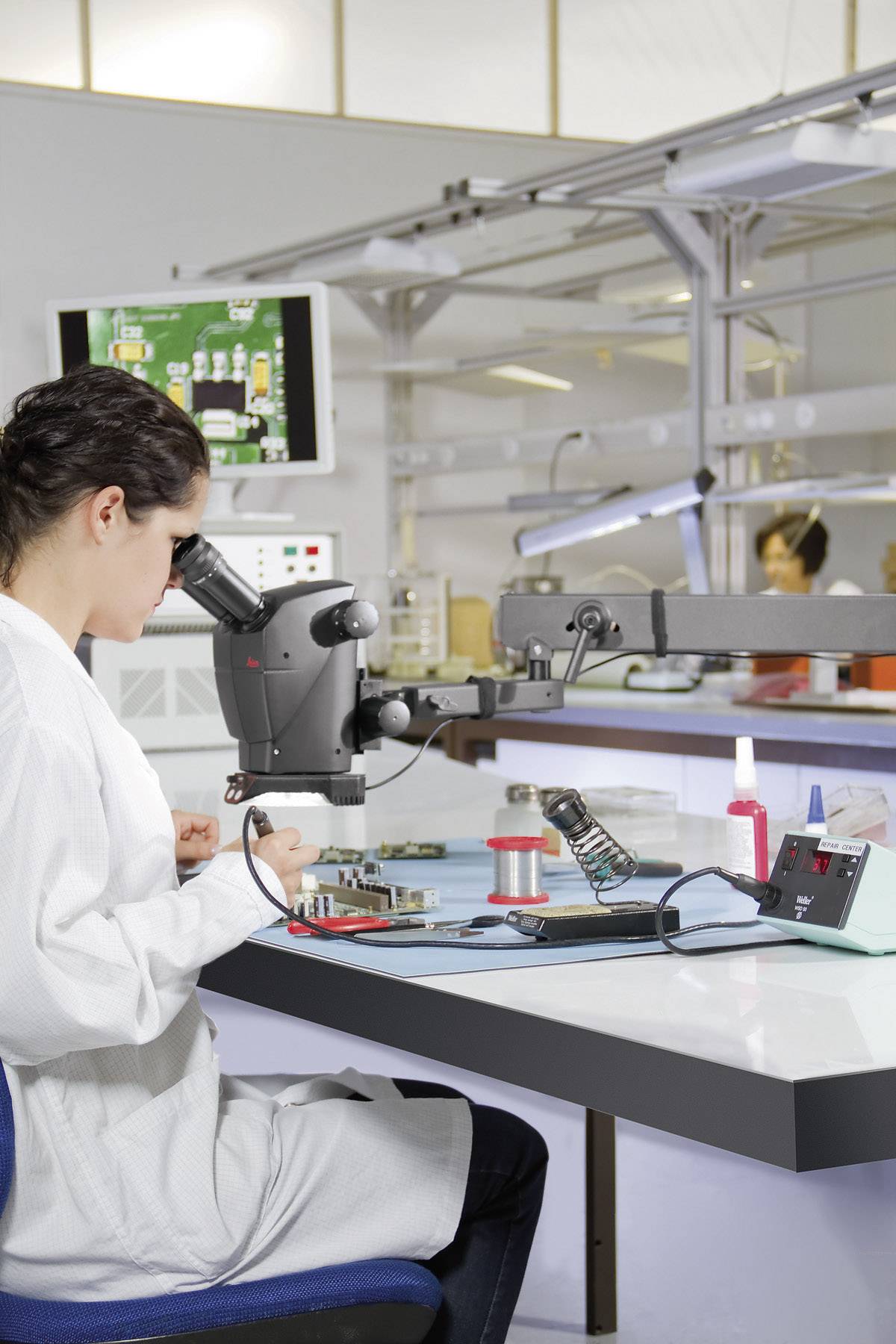 Egy laborköpenyt viselő személy egy mikroszkóppal egy elektronikai eszközön dolgozik egy laboratóriumban. A háttérben további laboratóriumi munkatársak és egy monitor látható.