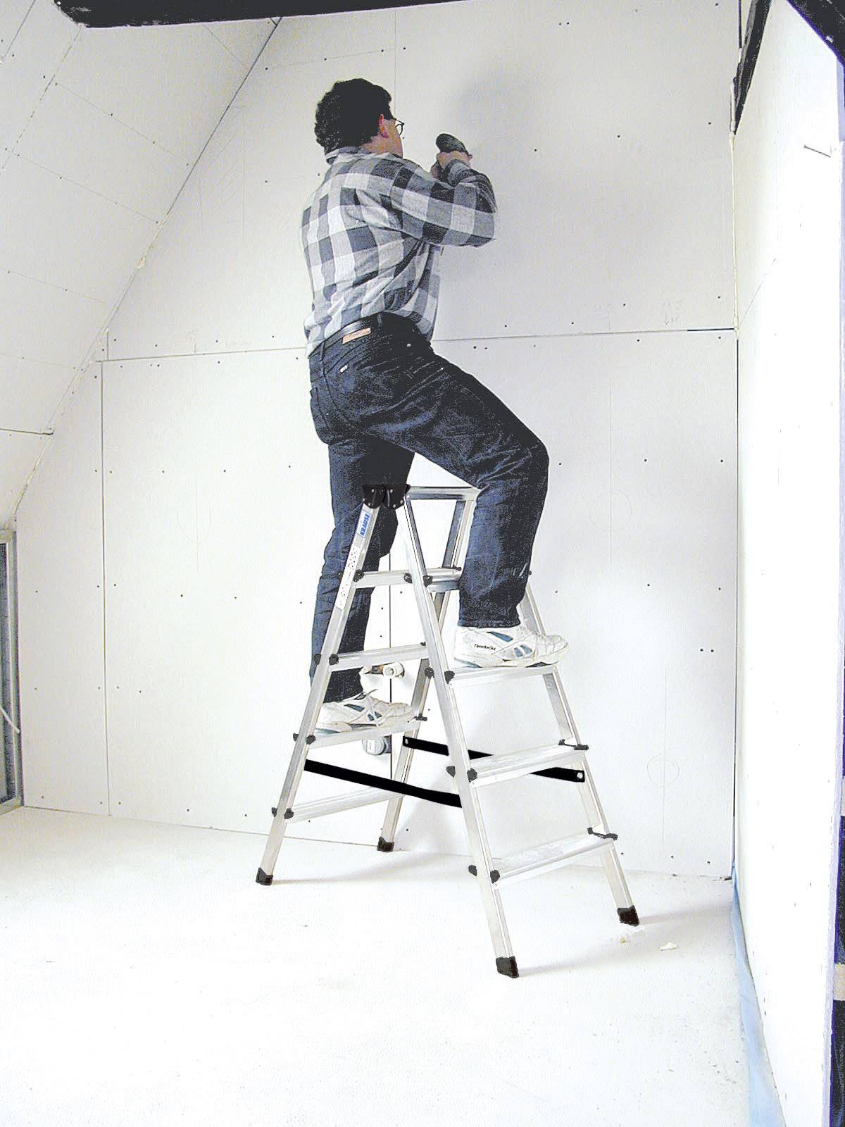 Egy kockás ingű és farmernadrágos ember áll egy létrán, és egy válaszfalat szerel egy helyiségben.