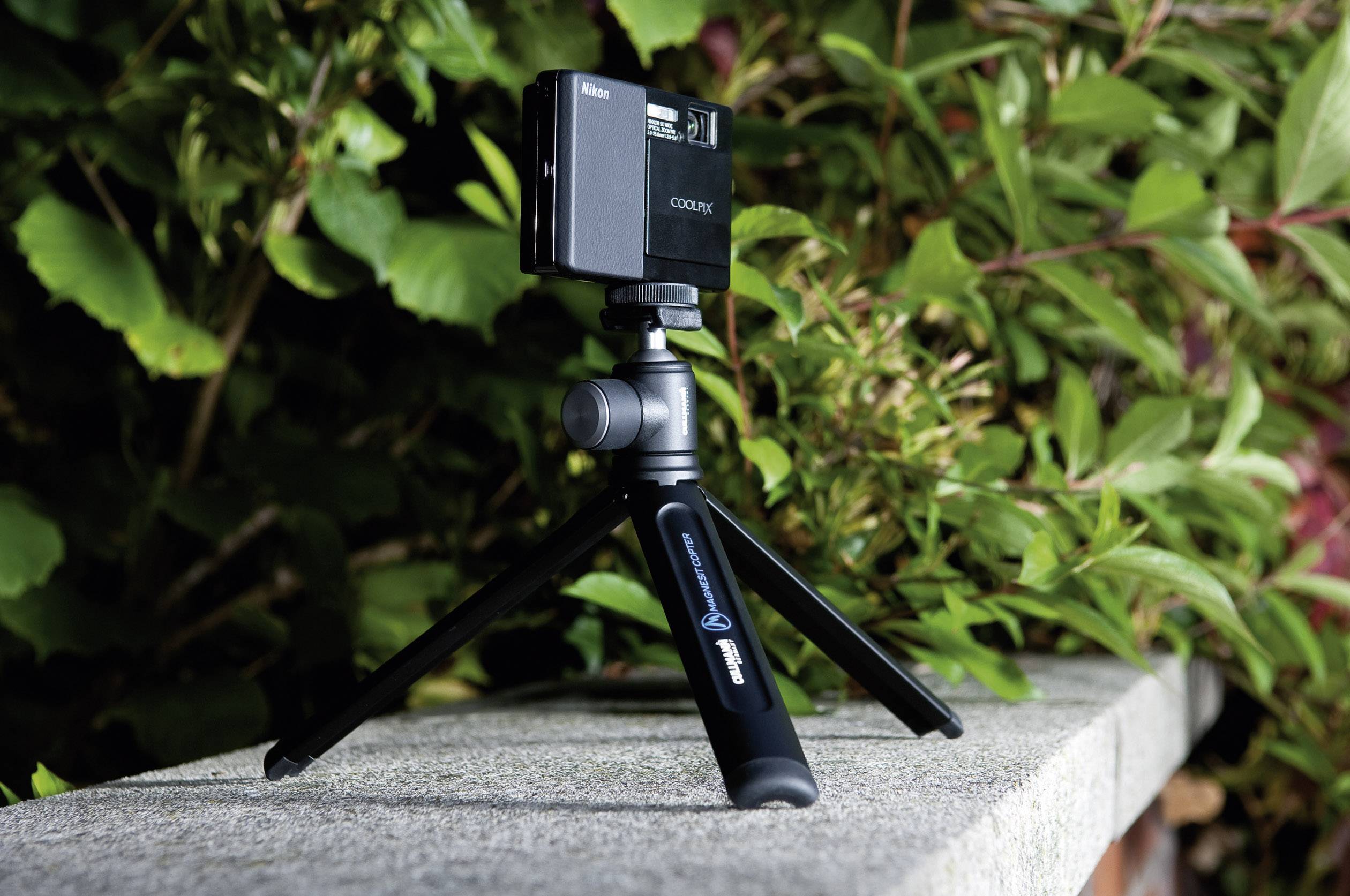 Egy fényképezőgép egy kompakt állványon egy kőfalon áll, zöld növények övezik a háttérben.