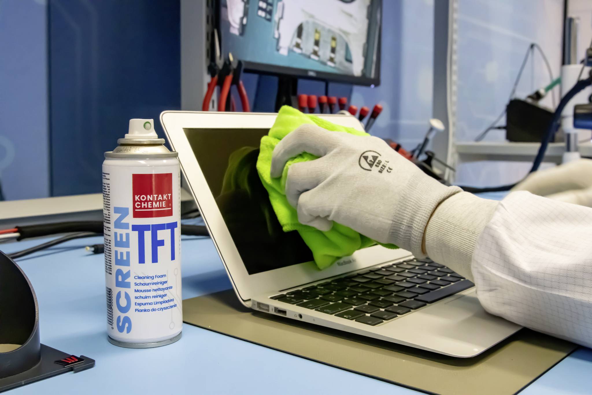 Una persona sta pulendo lo schermo di un laptop con un panno verde e un detergente per schermi in un ambiente tecnico.