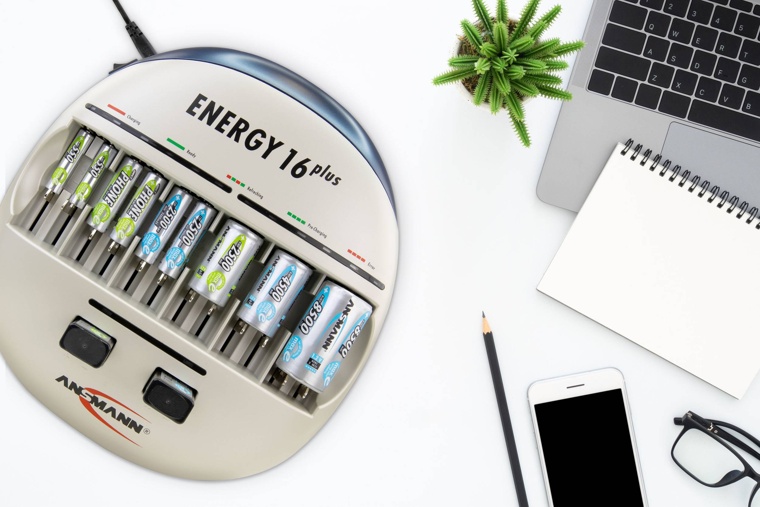 Caricabatterie con sei batterie AA inserite, accanto a un laptop, un quaderno, uno smartphone e una piccola pianta.