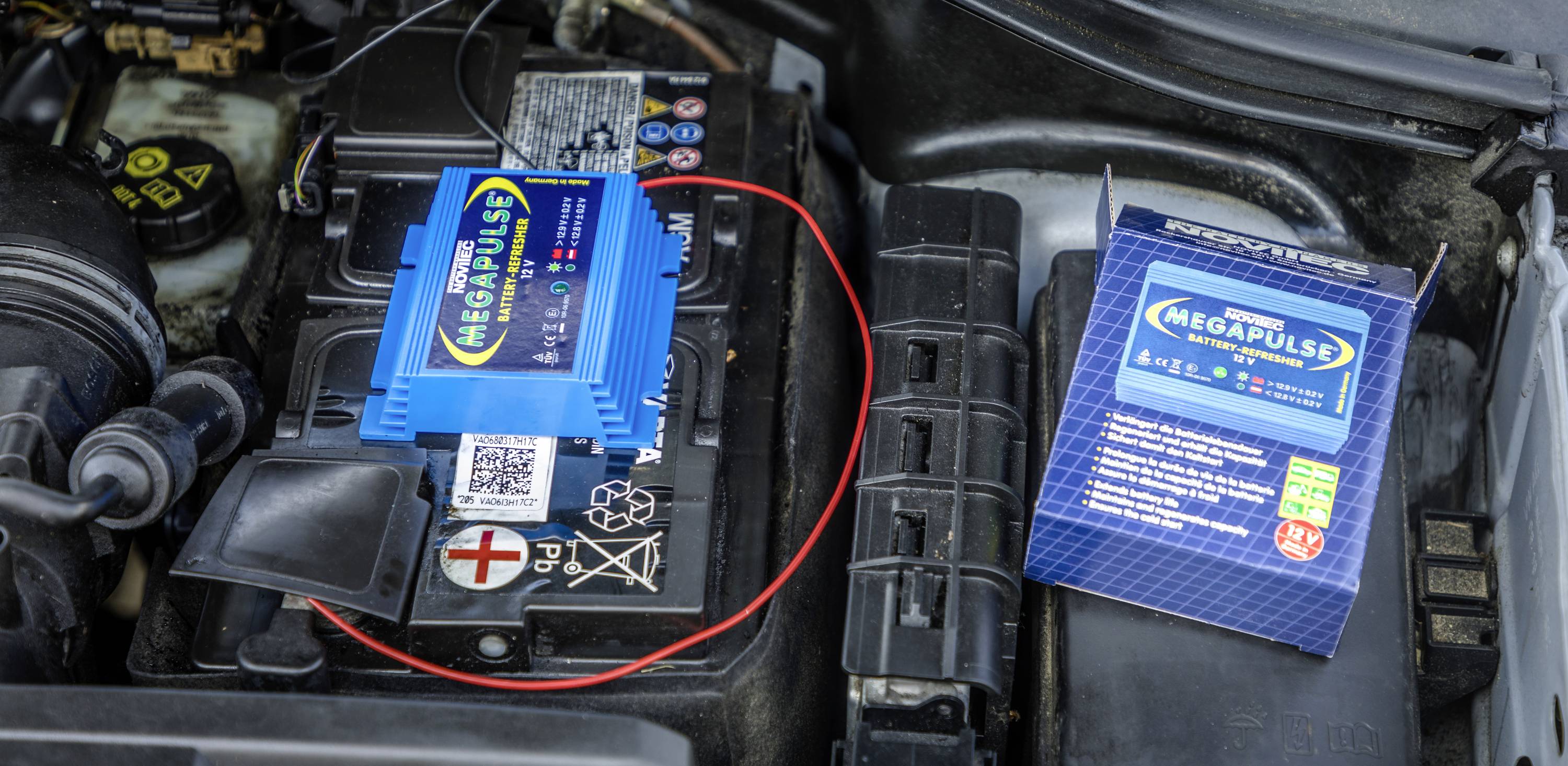 Un dispositivo Megapulse è collegato a una batteria per auto. Accanto a destra si trova la confezione. Il dispositivo serve per la manutenzione della batteria.