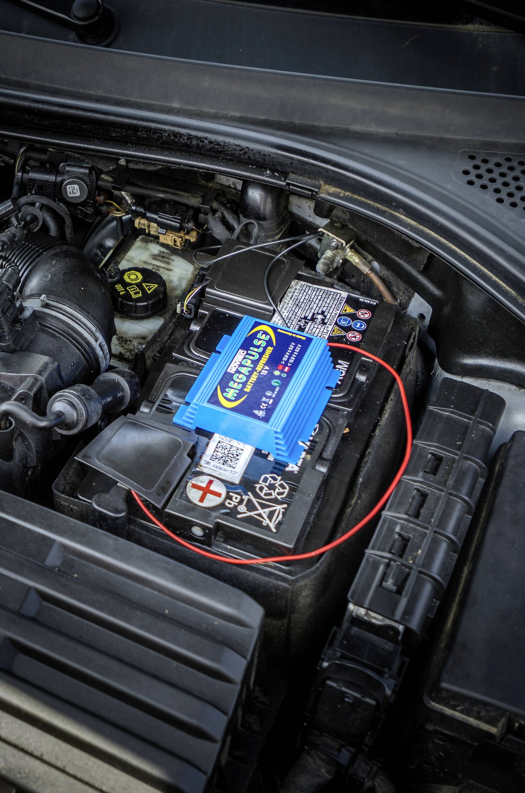 Batteria dell'auto nel vano motore con un dispositivo elettronico blu sopra di essa. Il dispositivo è collegato a un cavo rosso.