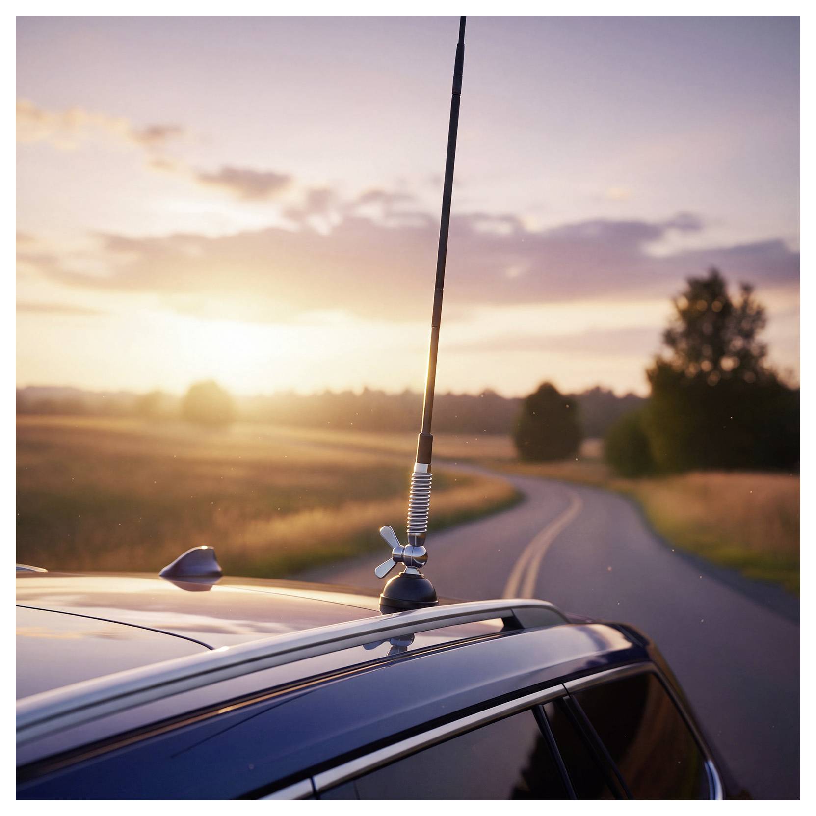 Tetto dell'auto con antenna su una strada rurale tortuosa al tramonto, con campi e alberi sullo sfondo.