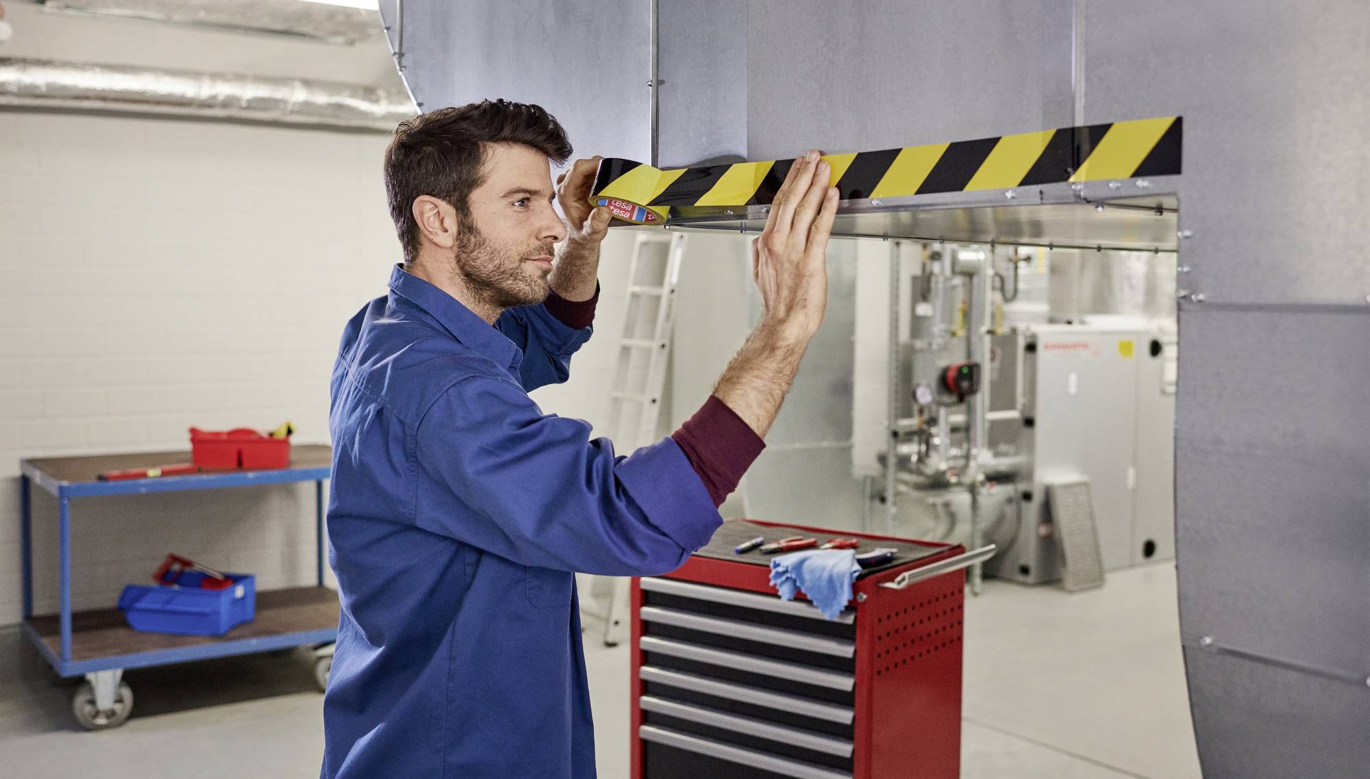 Un uomo con indumenti da lavoro blu sta applicando un nastro di avvertimento nero e giallo su un bordo metallico in un ambiente industriale. Sullo sfondo sono visibili attrezzi e macchinari.
