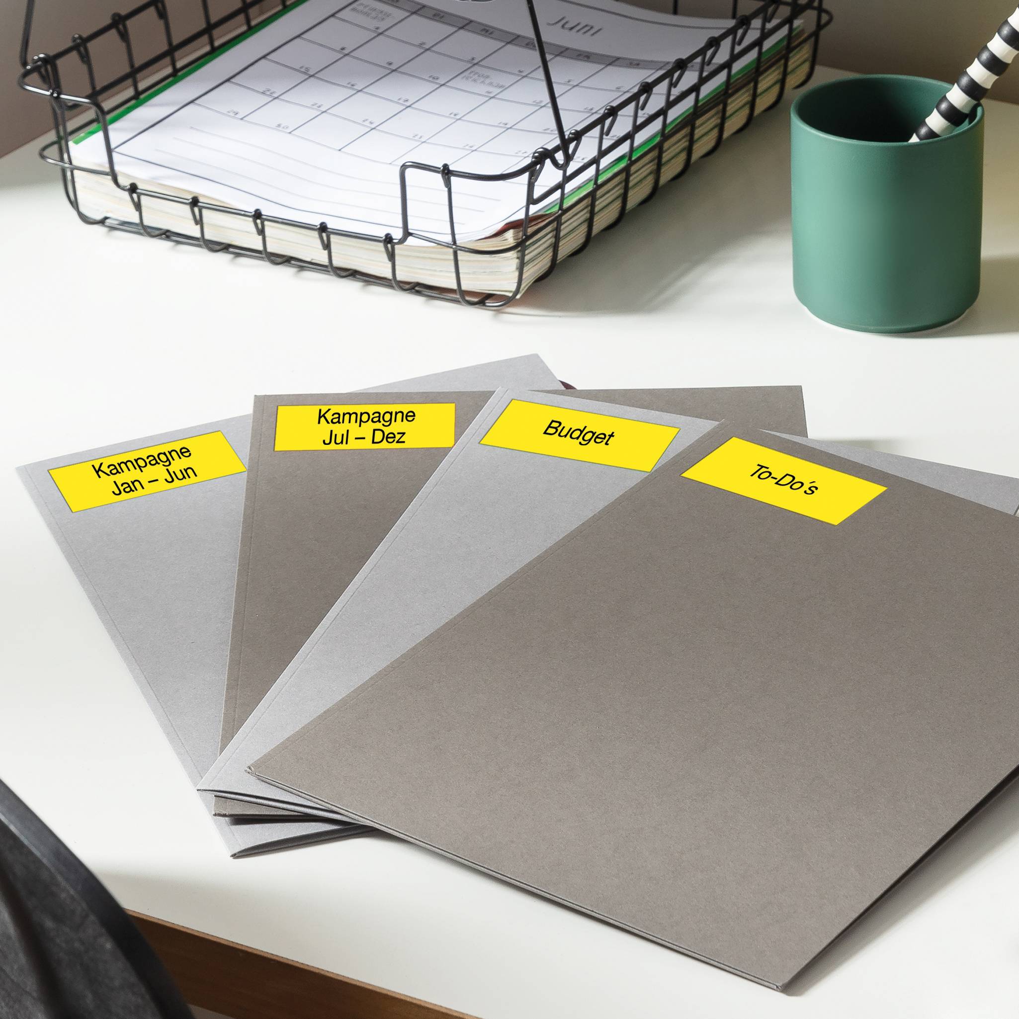 Quattro raccoglitori sono posizionati su una scrivania, etichettati con 'Campagna Gen - Giu', 'Campagna Lug - Dic', 'Budget' e 'To-Do'; sullo sfondo un calendario mensile da tavolo e un portapenne.