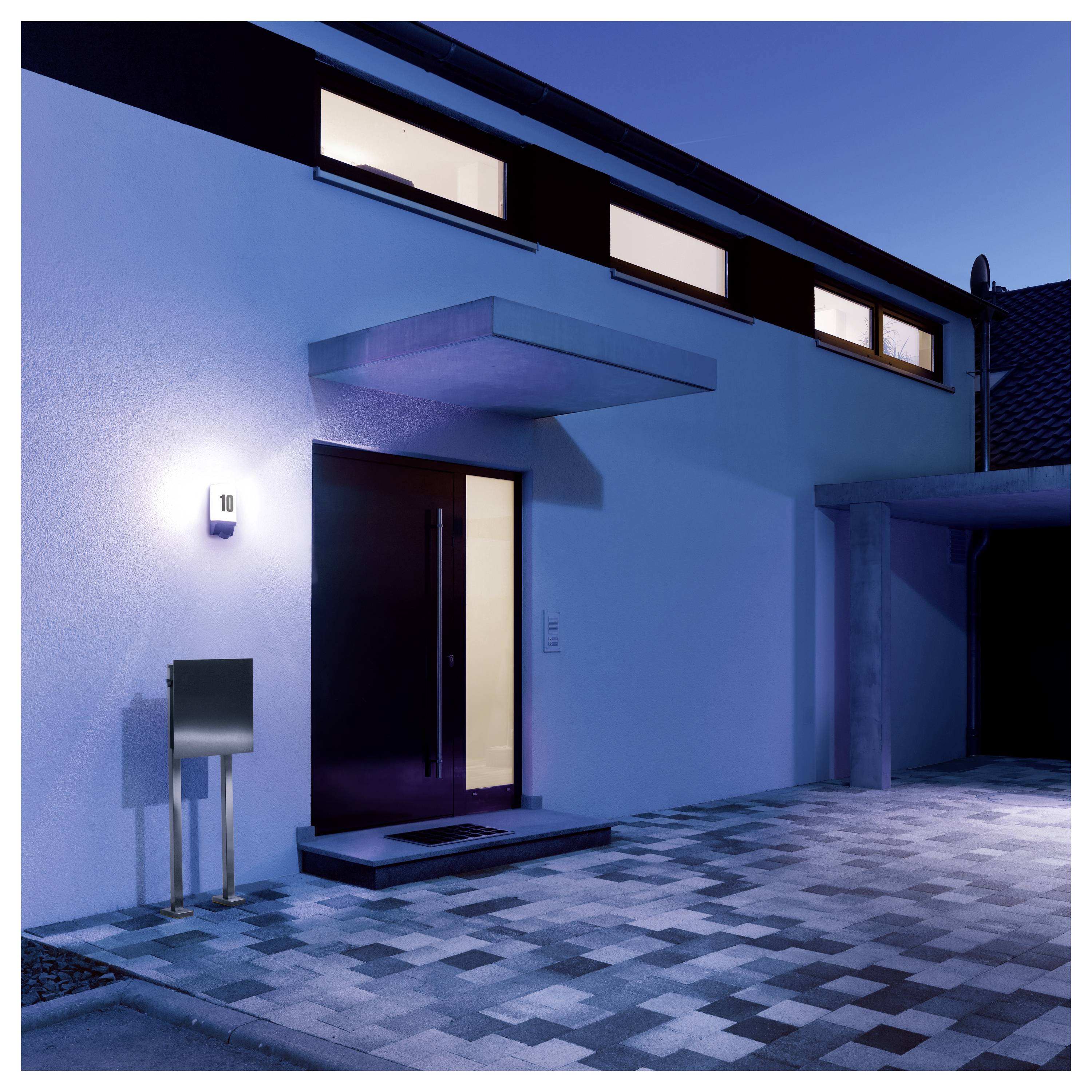 Ingresso di una casa moderna al tramonto con porta d'ingresso illuminata, numero civico 10 sulla parete, e un vialetto pavimentato che conduce al garage.