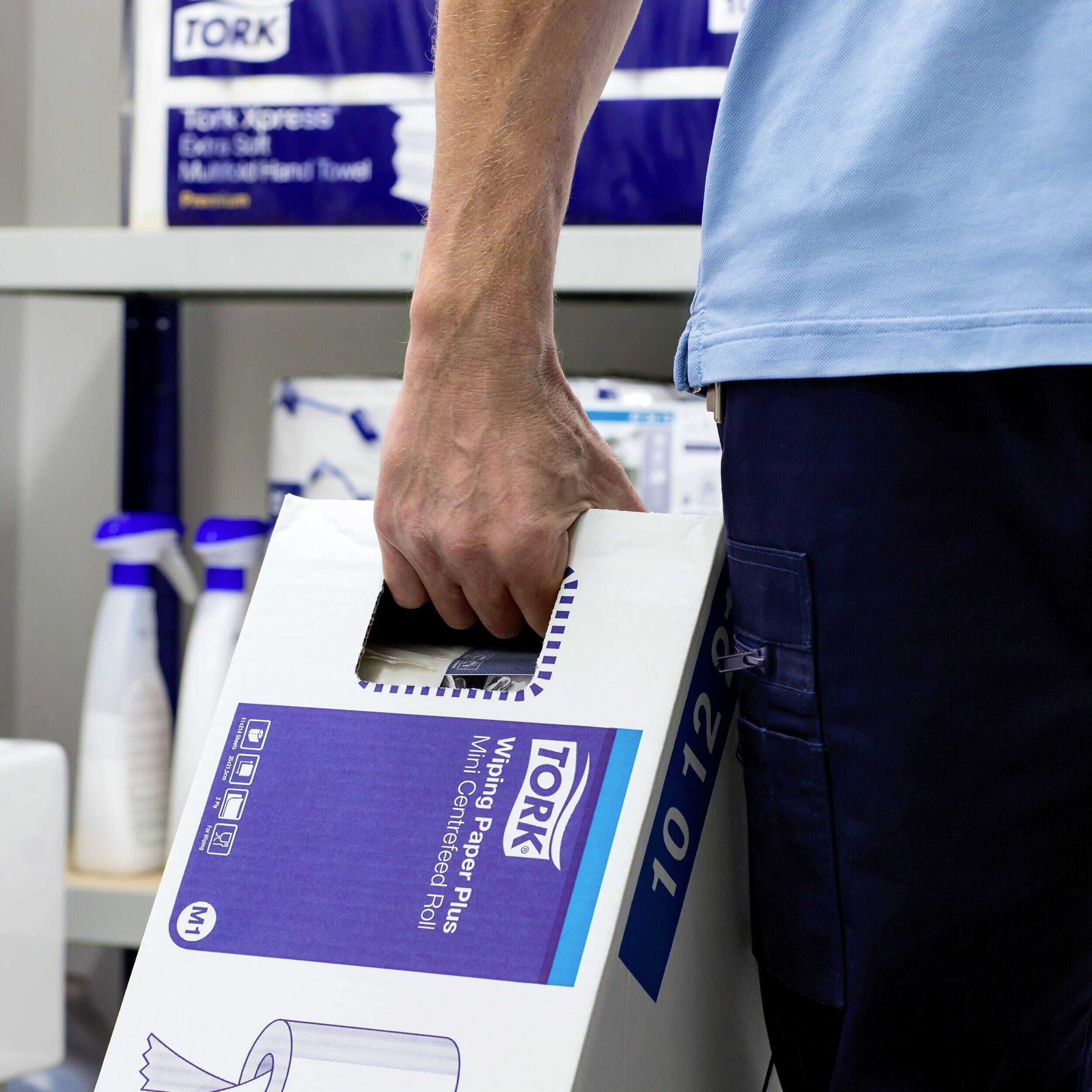 Una persona sta trasportando una scatola di fazzoletti di carta in un magazzino. Sullo sfondo ci sono altri prodotti per la pulizia e degli scaffali.