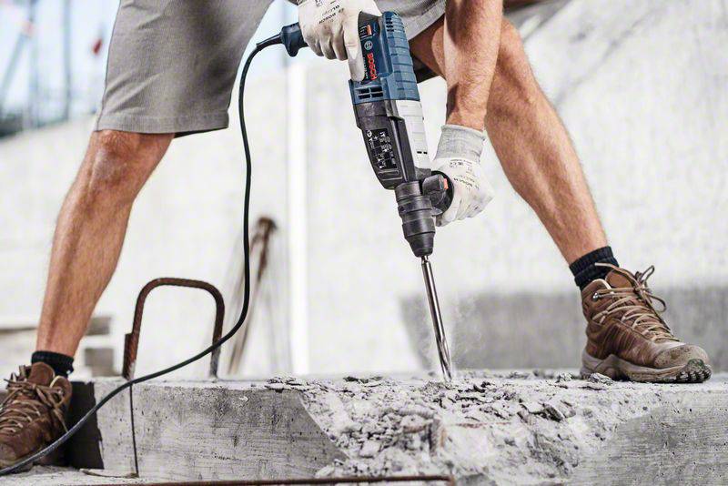 Una persona con guanti da lavoro utilizza un martello pneumatico per lavorare il calcestruzzo. Sullo sfondo sono visibili ponteggi edili.