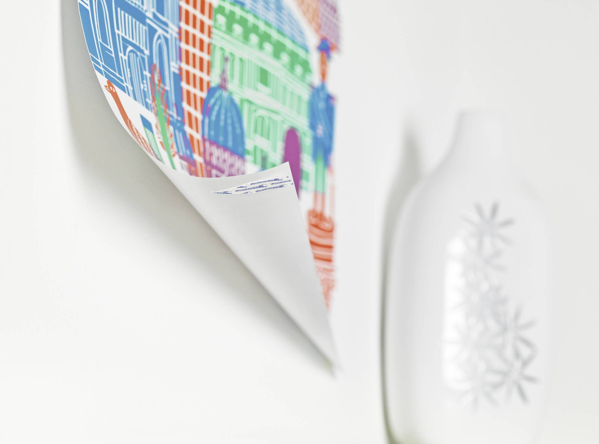 Un poster artisticamente realizzato, colorato, con edifici stilizzati e forme, è appeso a una parete bianca accanto a un vaso bianco.