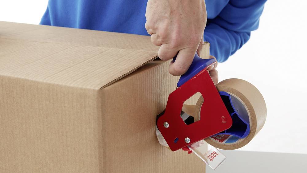 Una persona chiude una scatola con nastro adesivo utilizzando un dispenser rosso.