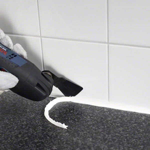 Una mano impugna un utensile elettrico con un accessorio per spatola al fine di rimuovere il sigillante bianco dall'angolo di una superficie piastrellata.