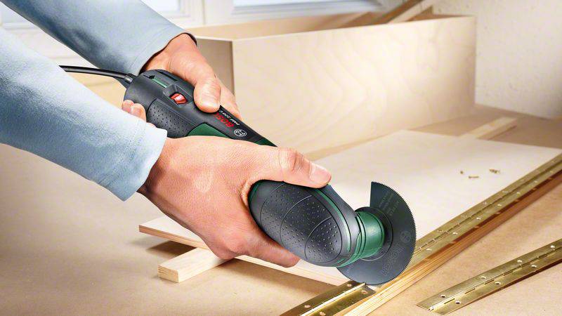 Una persona taglia con precisione un pezzo di legno utilizzando un utensile multifunzione elettrico. Viti e listelli di legno sono posizionati accanto.