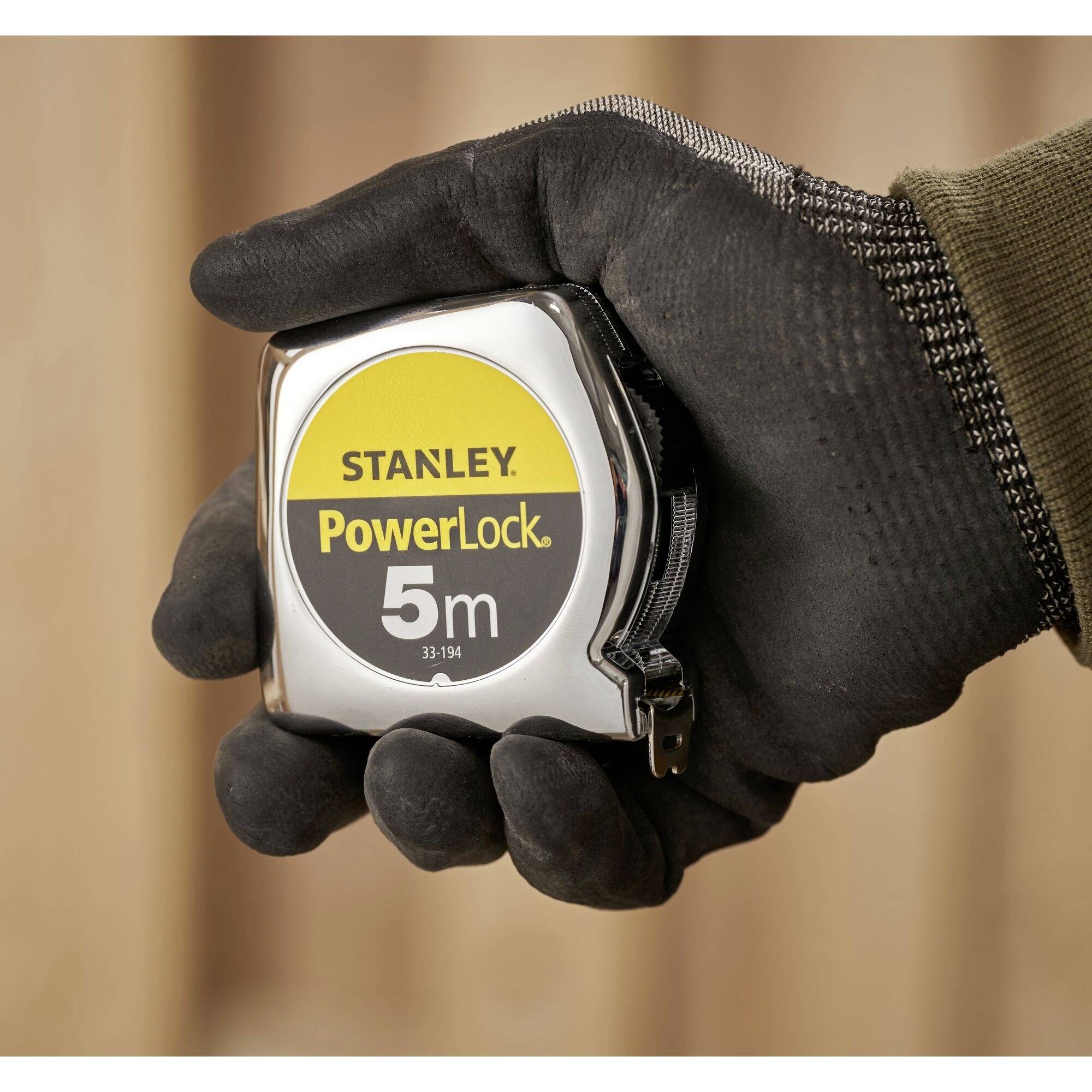 Una mano con un guanto tiene un metro a nastro Stanley PowerLock da 5 metri, con involucro giallo e argentato.