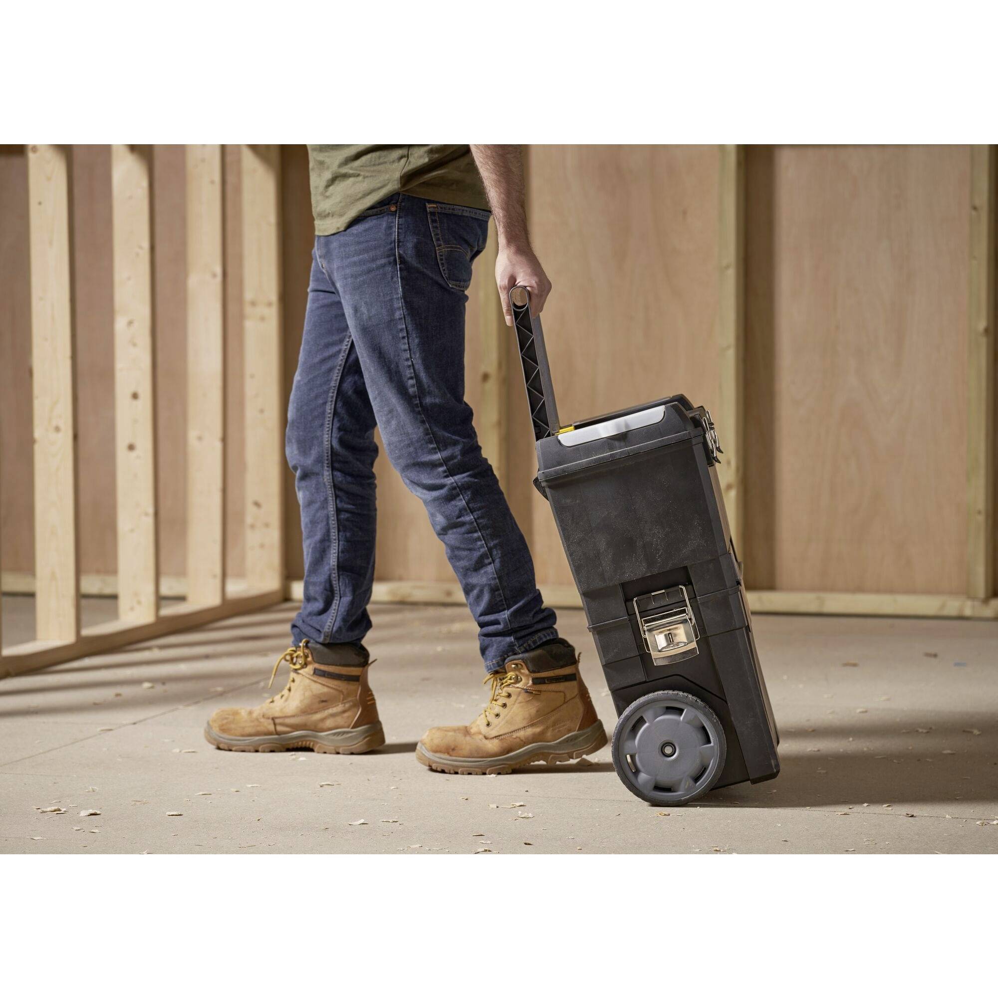 Una persona trascina una scatola degli attrezzi nera su ruote attraverso un pavimento di legno non finito. Indossa jeans e stivali da lavoro.