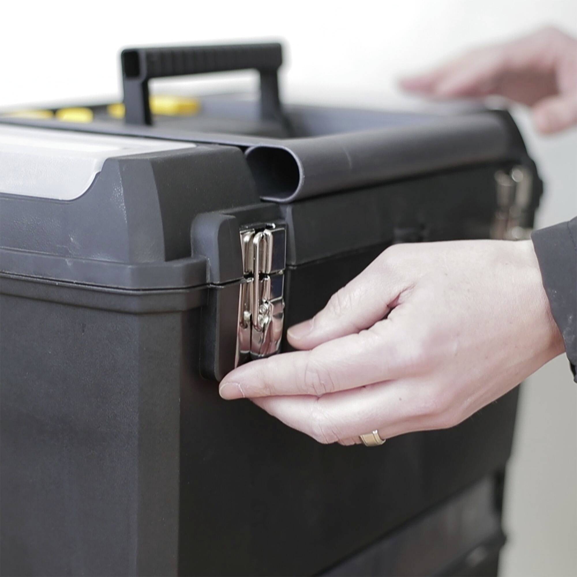 Una mano apre il fermo metallico di un cassetta degli attrezzi nera. La cassetta ha un manico per il trasporto e bottoni gialli visibili sul coperchio.